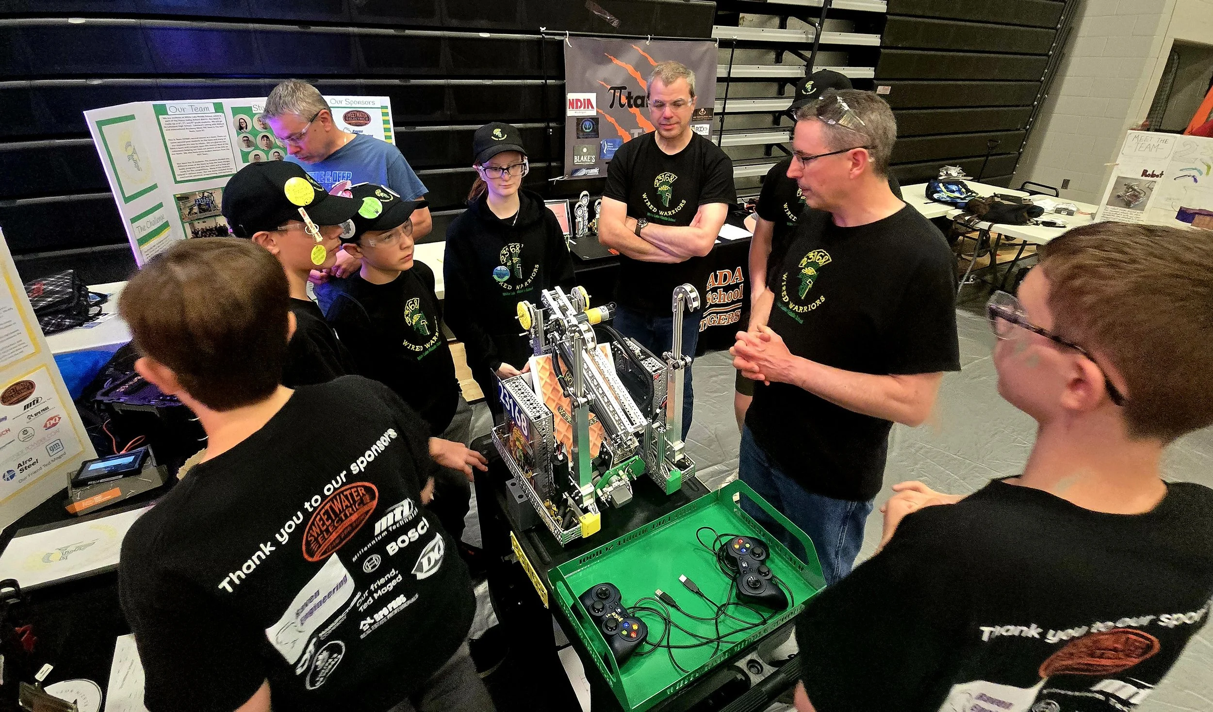 A group of students and mentors gathered around a robotics project at a science fair, with posters and tables in the background.