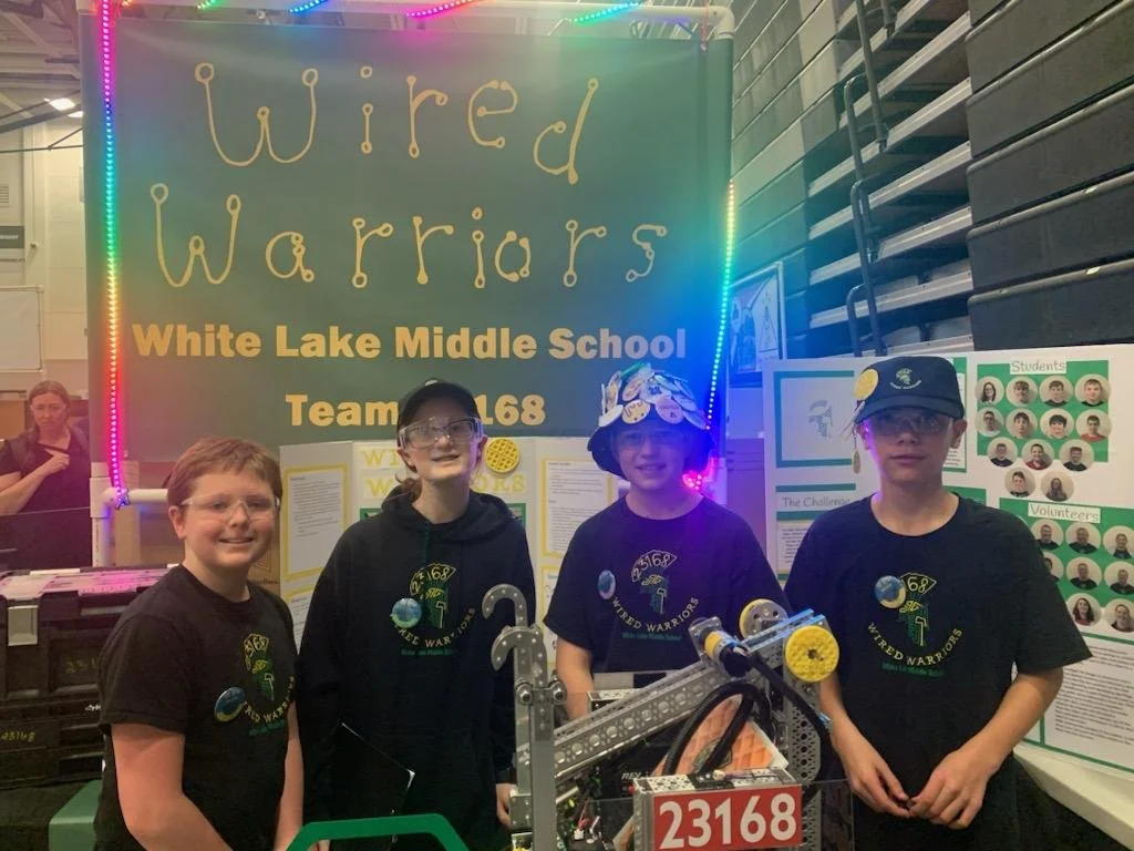 Four students wearing black T-shirts with a planet and robot logo, some with glasses, standing in front of a sign that reads "Wired Warriors" and "White Lake Middle School Team 23168." They are in a gymnasium with colorful lights, and a display with 