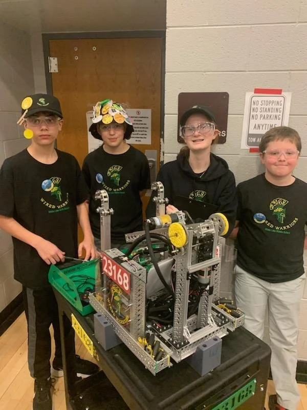 Four children, wearing matching black T-shirts with a robotics logo, stand behind a robot on a table. The robot is made of metal, with visible gears and wires, and has a red number 23168 on it. They are in a room with a wooden door and some signs on 