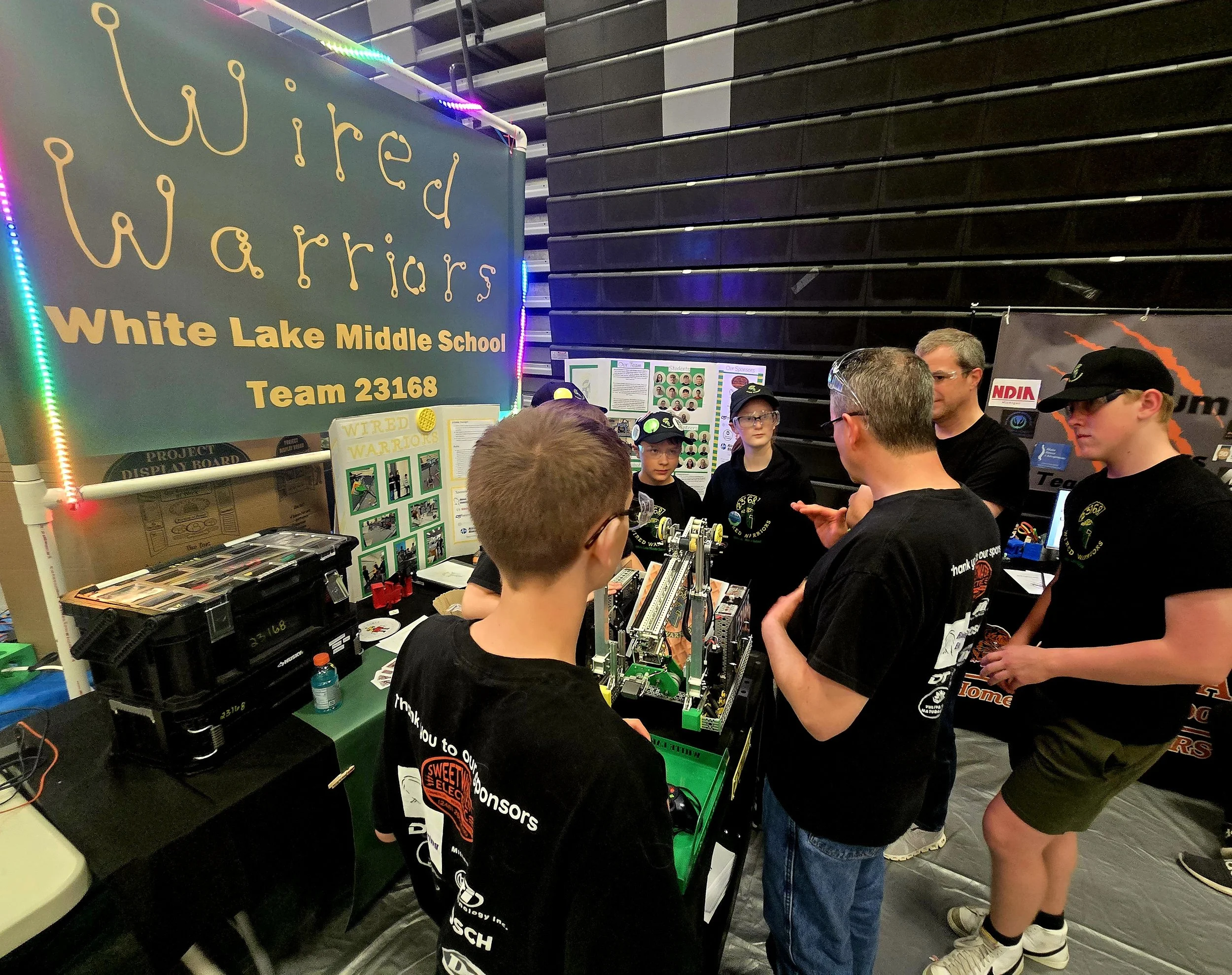 Group of middle school students and two adults gathered around a robotics project at an indoor science fair or competition, with a large blue sign reading "Wired Warriors, White Lake Middle School, Team 23168" in the background.