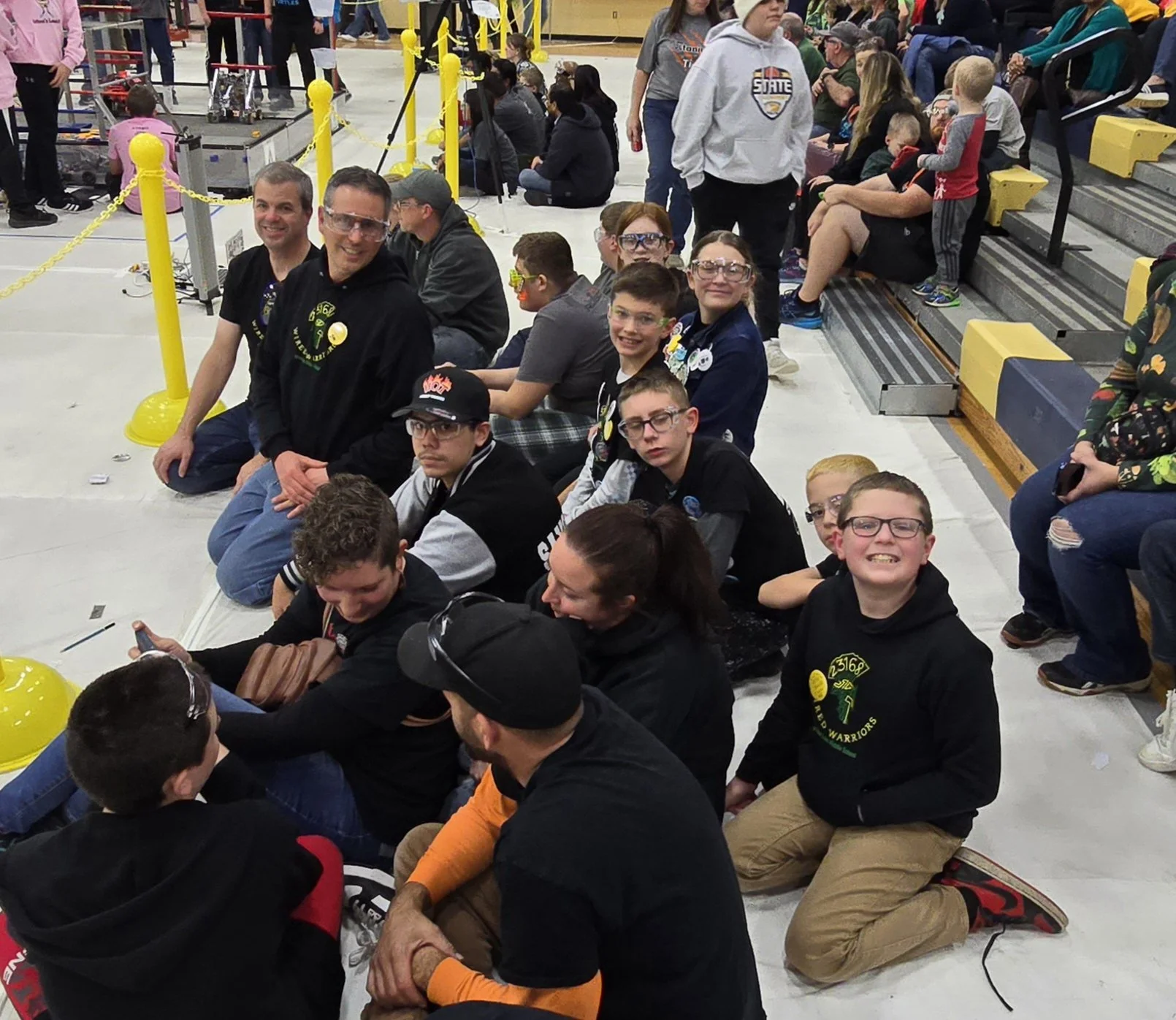 A group of people, including children and adults, seated on the floor and benches inside a large indoor space, possibly a gymnasium, watching a robotics competition with robots and safety barriers in the background.