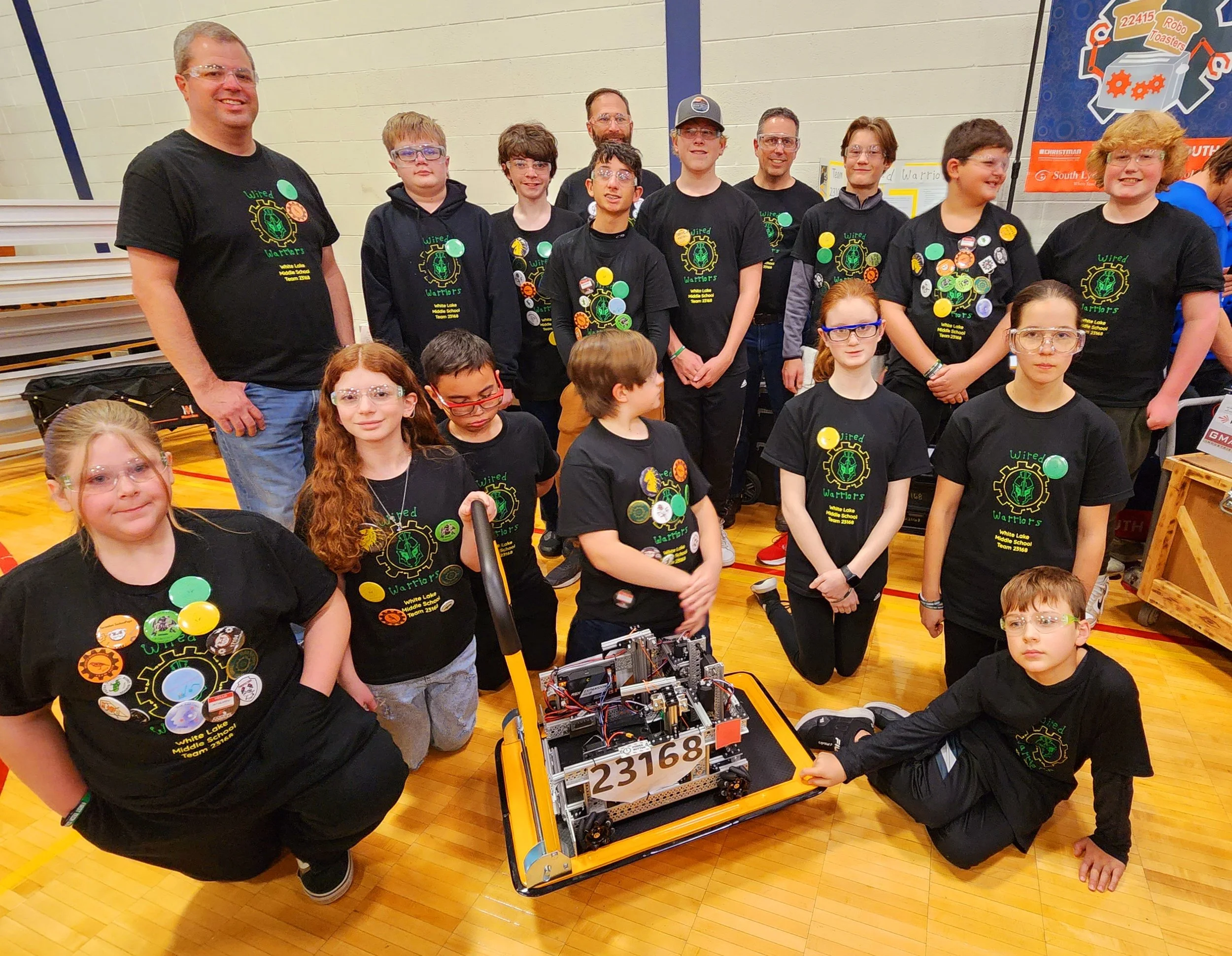 Group of children and adults in matching black shirts with a green gear logo, gathered around a robot with the number 23168 inside a gymnasium, some kneeling, some standing, posing for a team photo.