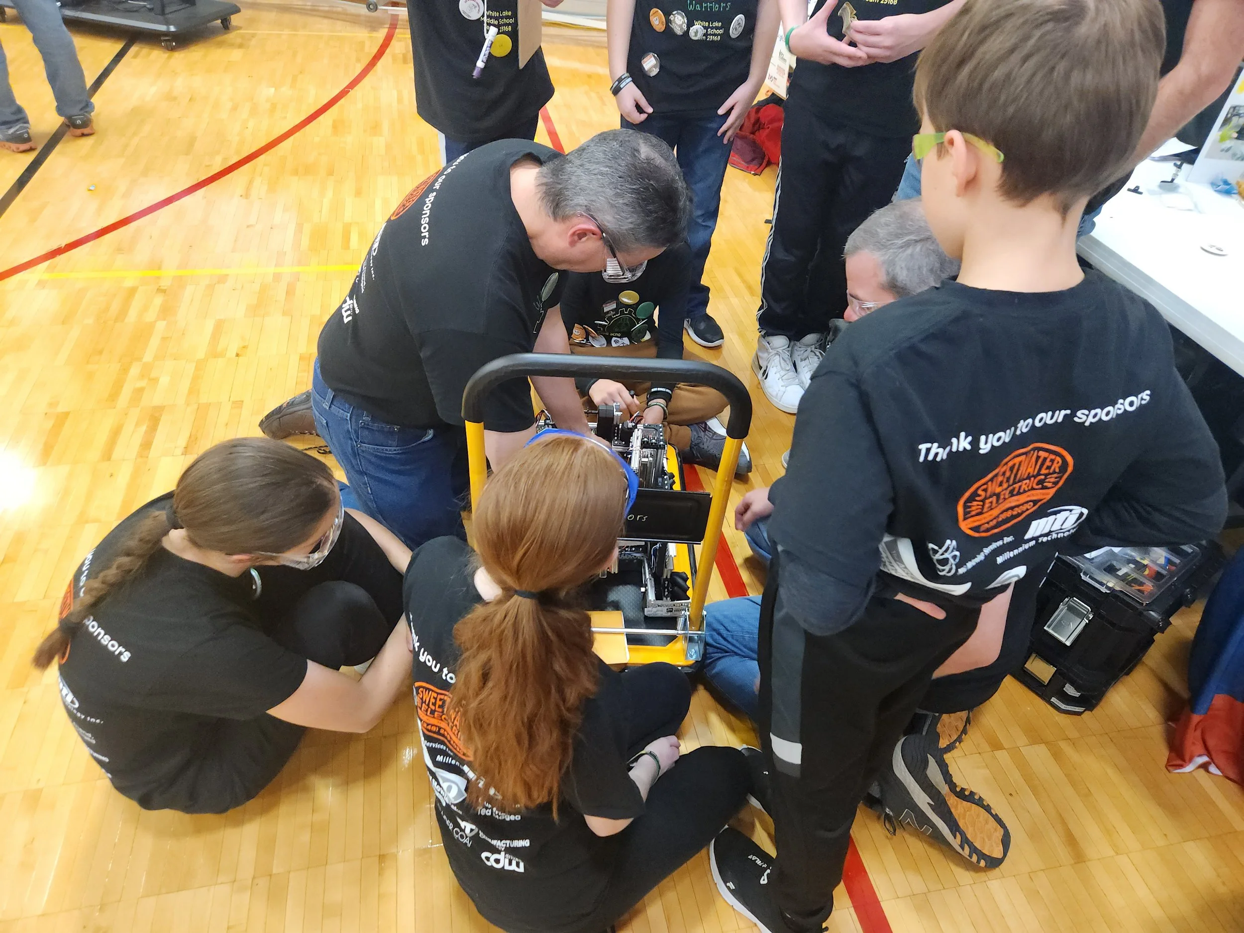 Group of students and an adult gather around a robot on a gymnasium floor, working together on a project.