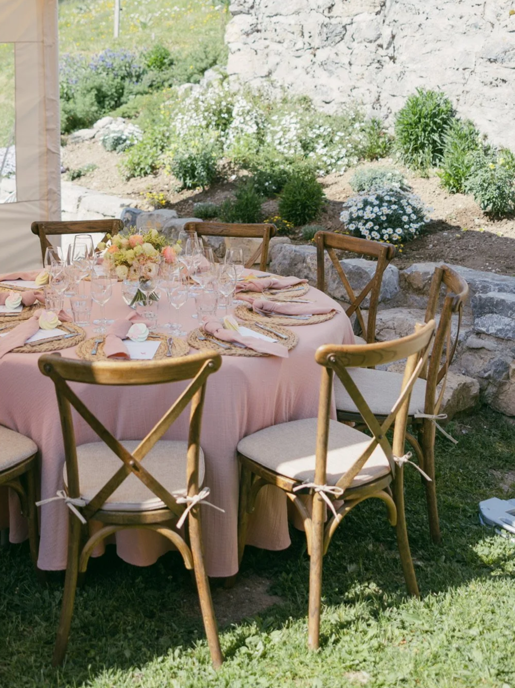 Table de banquet décorée avec une nappe rose, des assiettes en rotin, des verres à vin et des couverts, entourée de chaises en bois avec des coussins beige, dans un jardin avec des fleurs et des plantes