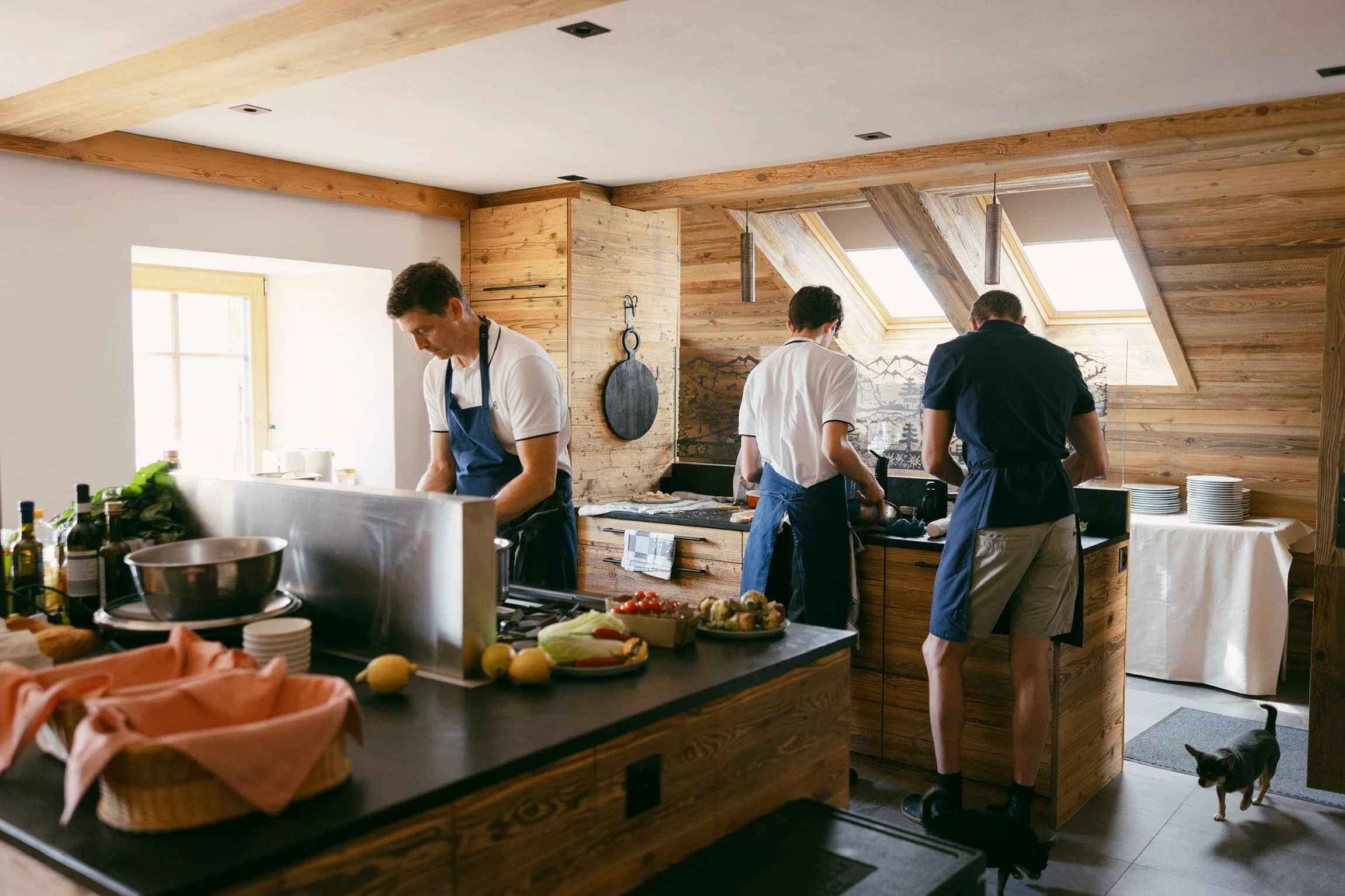 Cuisiniers préparant un repas dans une cuisine en bois lors d'un événement ou d'un cours de cuisine, avec de la nourriture et des ustensiles sur le comptoir.