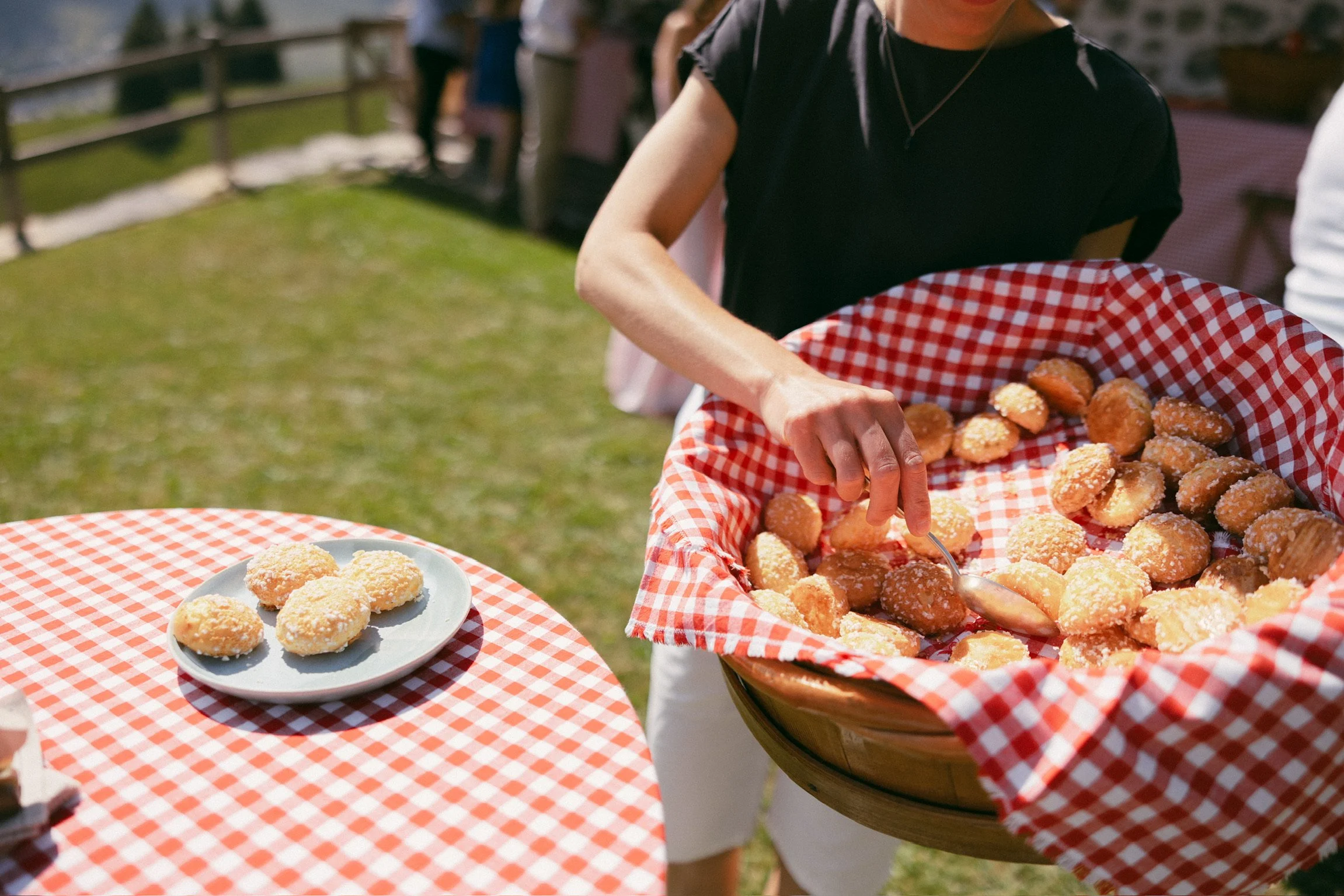 Une personne sert des chouquettes, petites friandises sucrées, dans un panier recouvert d'une nappe à carreaux rouges et blancs lors d'une fête en extérieur.