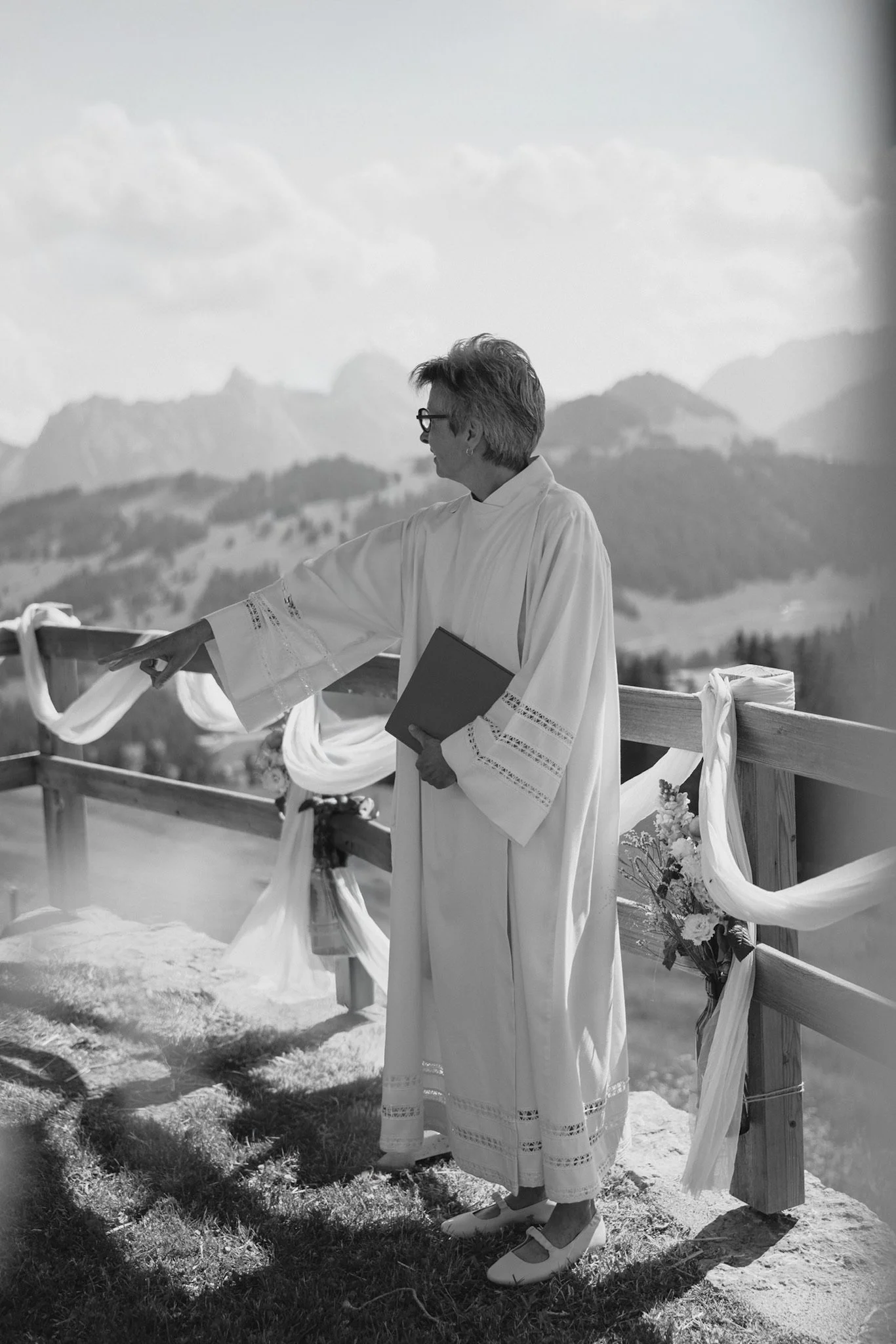 Une maitre de cérémonie tenant un livre, se tient devant une barrière en bois décorée avec des drapes et des fleurs, avec des montagnes en arrière-plan.