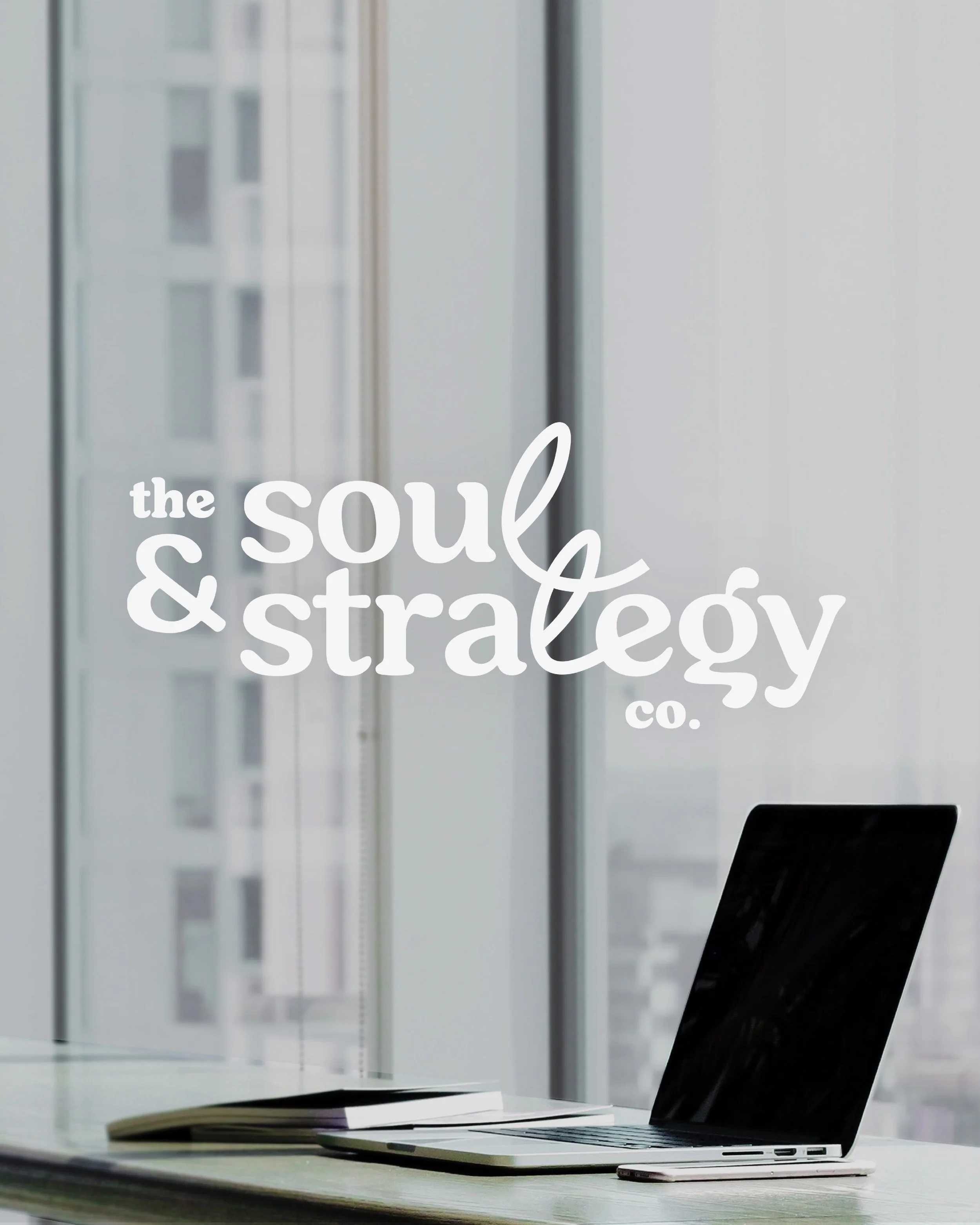 A modern workspace with a MacBook laptop, iPhone, and a closed book or document on a desk, with window blinds in the background and a logo overlay that says "The Soul & Strategy Co".