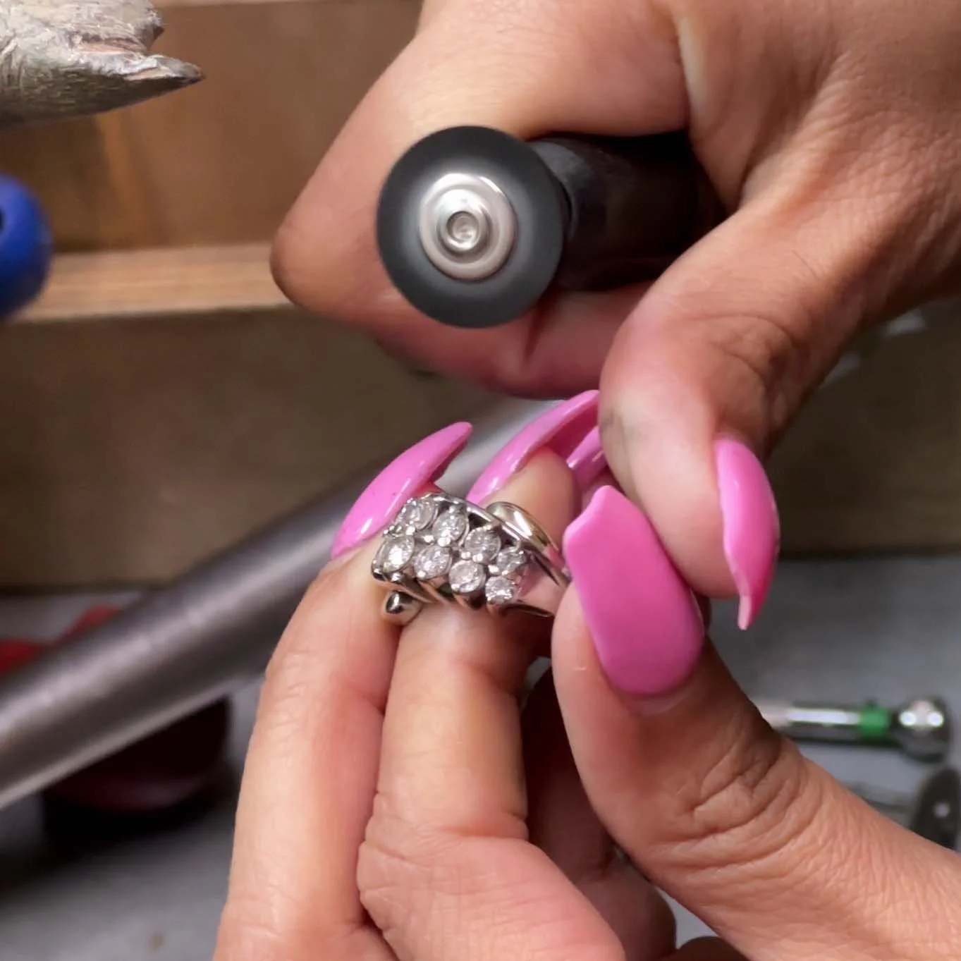 Close-up of a jewelry repair process: a person with pink painted nails holds a ring with diamonds while another hand uses a rotary tool to work on it.