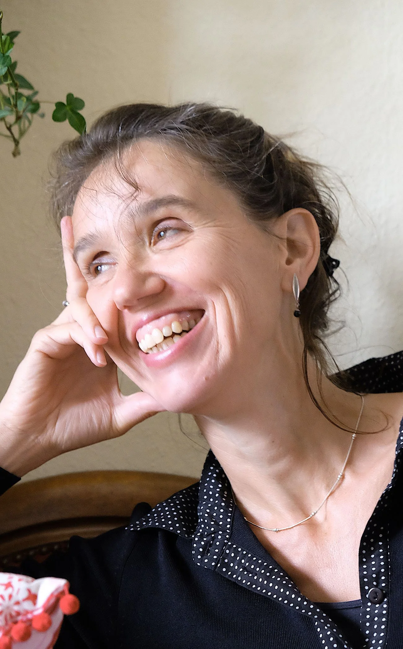 Eine lachende Frau mit Kurzhaarfrisur, die sich die Hand ans Gesicht hält. Sie trägt Ohrringe, eine zarte Halskette und ein schwarzes Oberteil mit weißen Punkten.