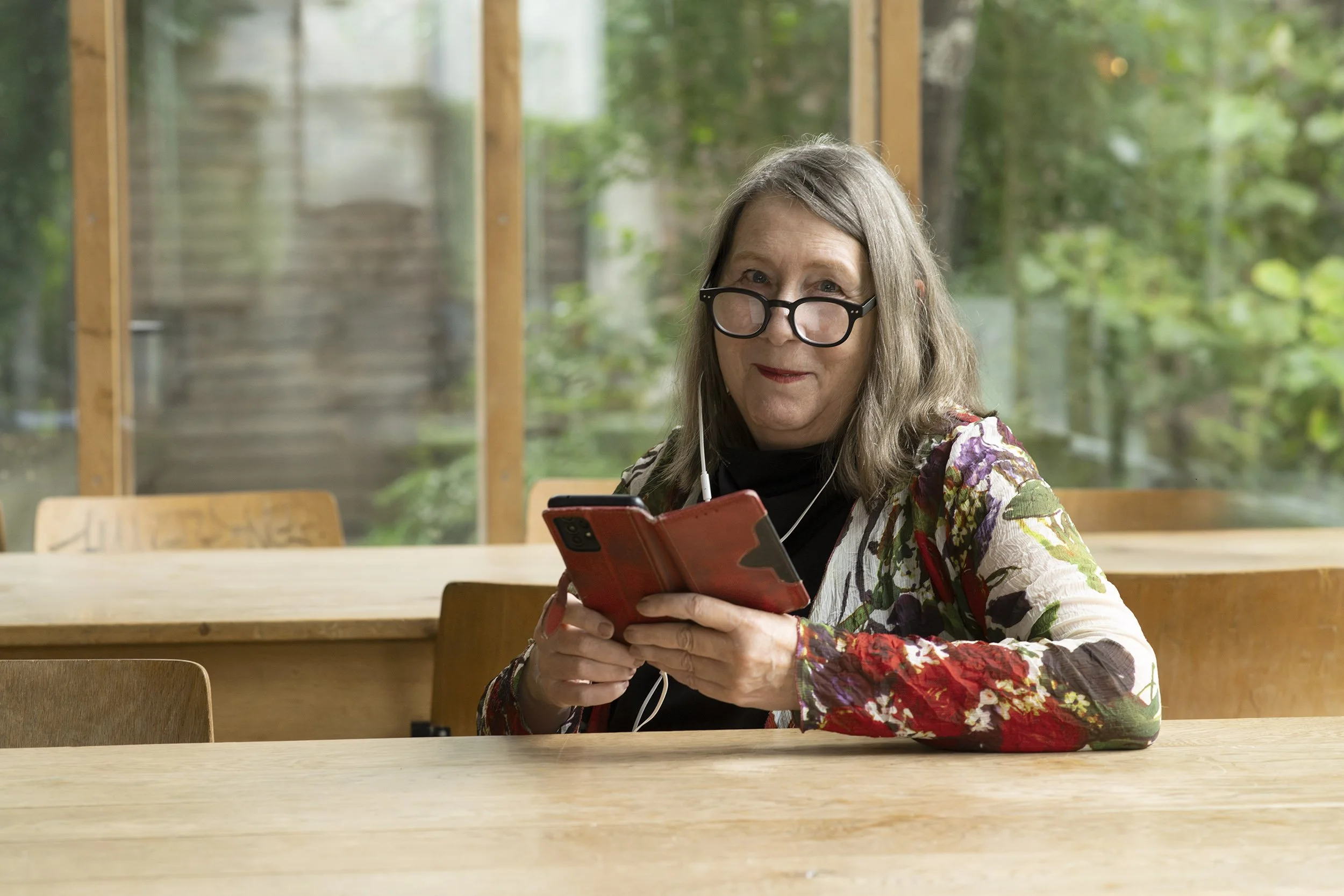 Ältere Frau mit Brille, sitzt an einem Holztisch, schaut in ihr Smartphone, hört Musik über Kopfhörer, im Hintergrund große Fenster mit Blick nach draußen auf einen Garten.