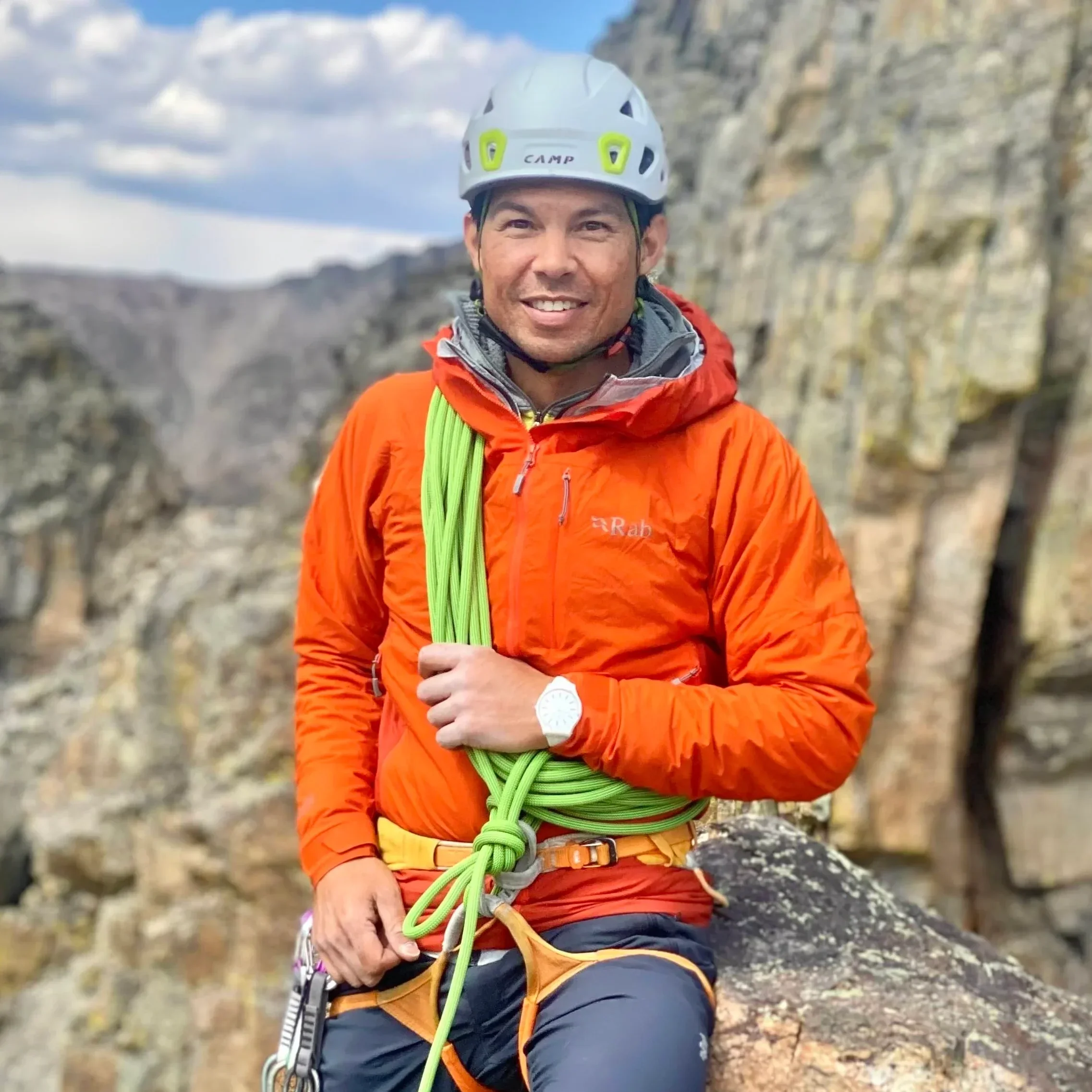 A man in climbing gear, wearing an orange jacket and a white helmet, is smiling outdoors with rocky cliffs in the background.