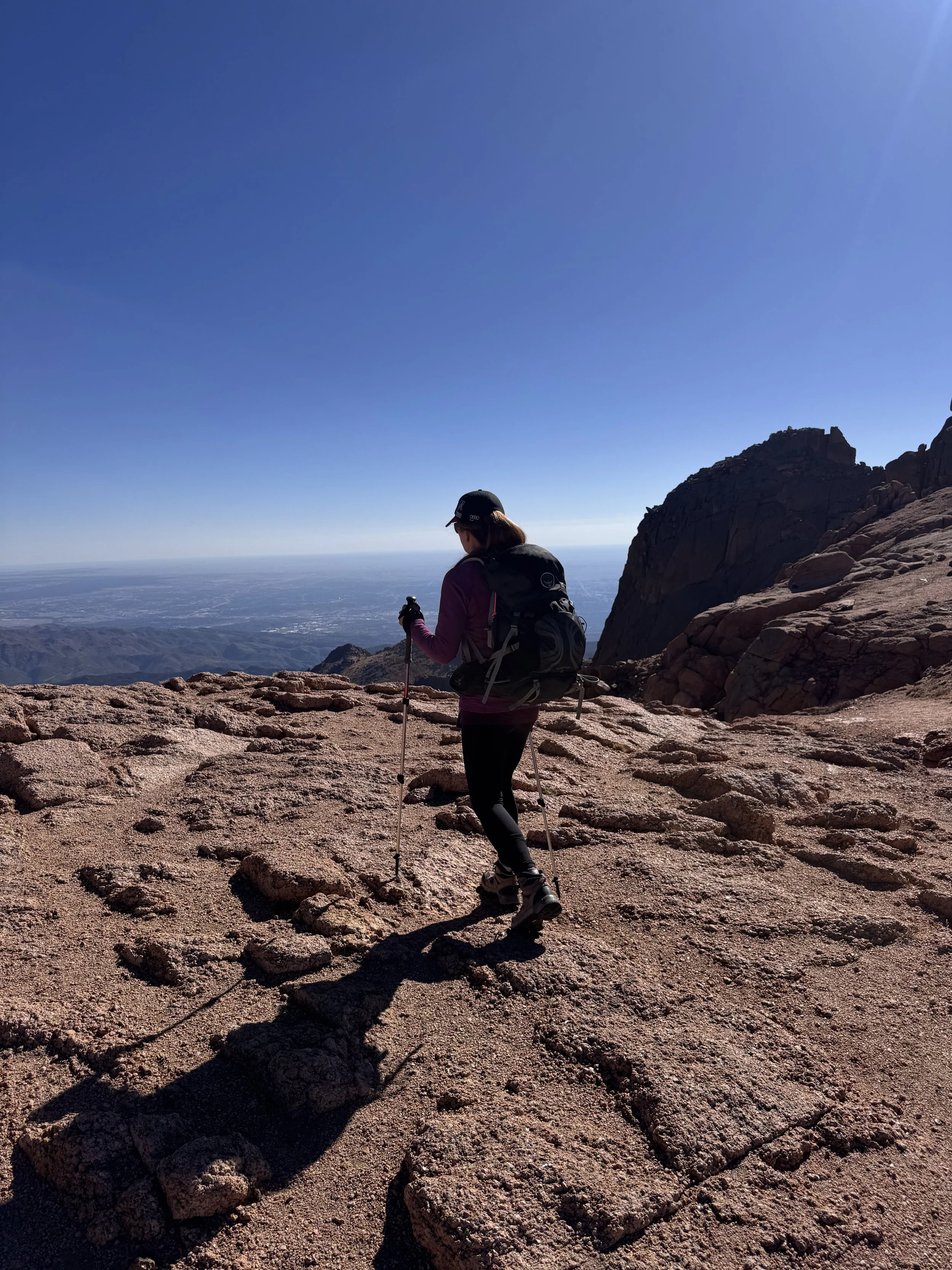 A hiker in outdoor gear climbing rocky terrain on a mountain with a scenic view and clear blue sky.