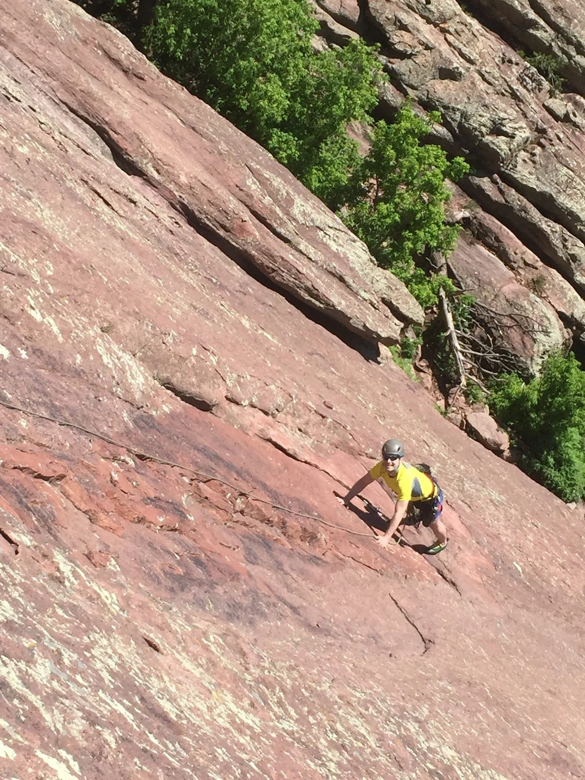 A person wearing a yellow shirt, helmet, and climbing gear is rock climbing on a steep, reddish-brown rock face with green trees in the background.