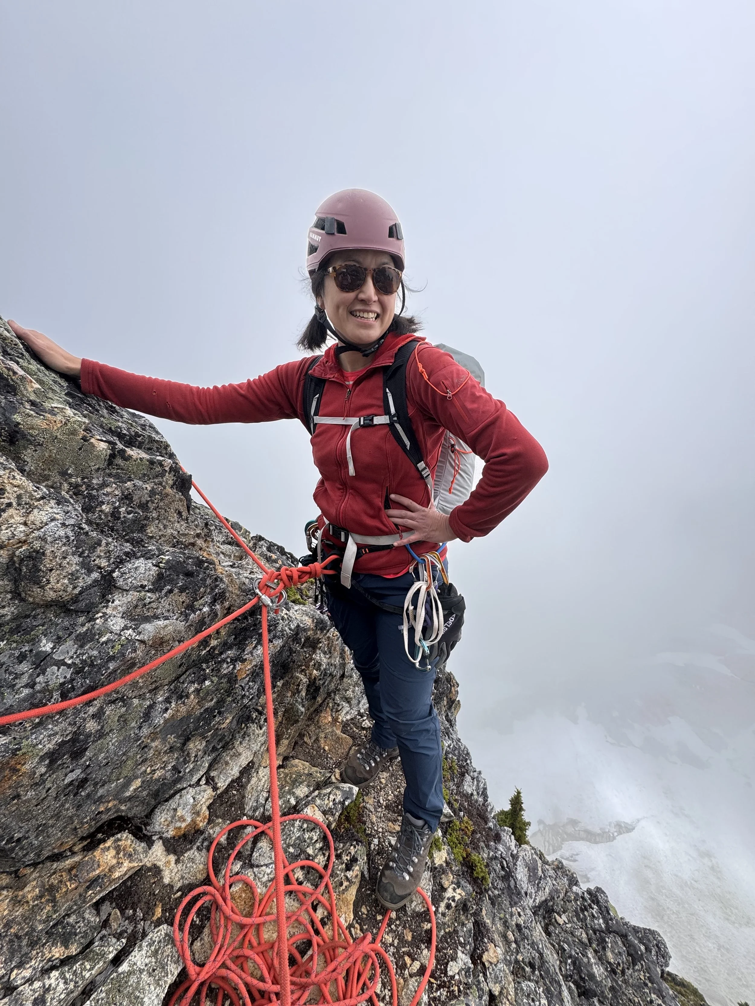A woman mountain climbing on a rocky ledge, wearing a pink helmet, sunglasses, a red jacket, and climbing gear, with a foggy background.