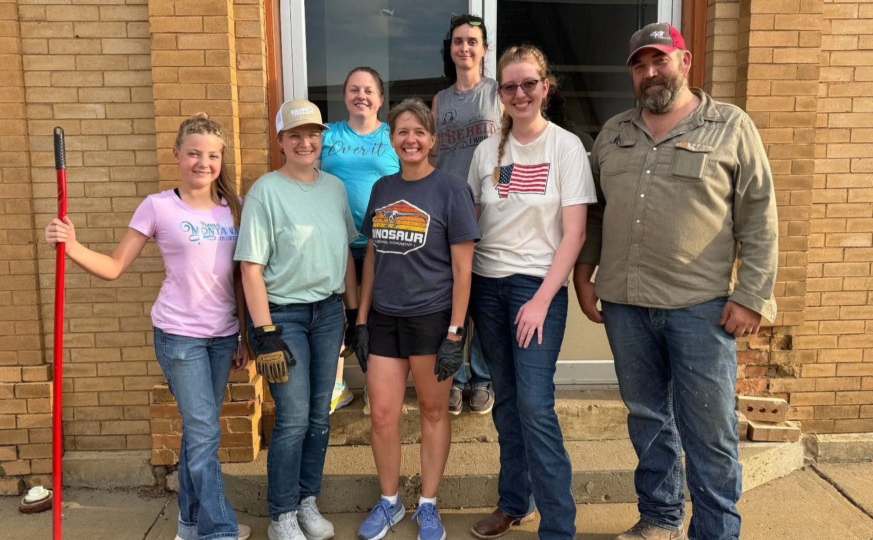 Group of eight people standing outside in front of a brick building, smiling at the camera. They are dressed casually, some wearing gloves, with one person holding a cleaning tool, volunteer activity for a cleanup.