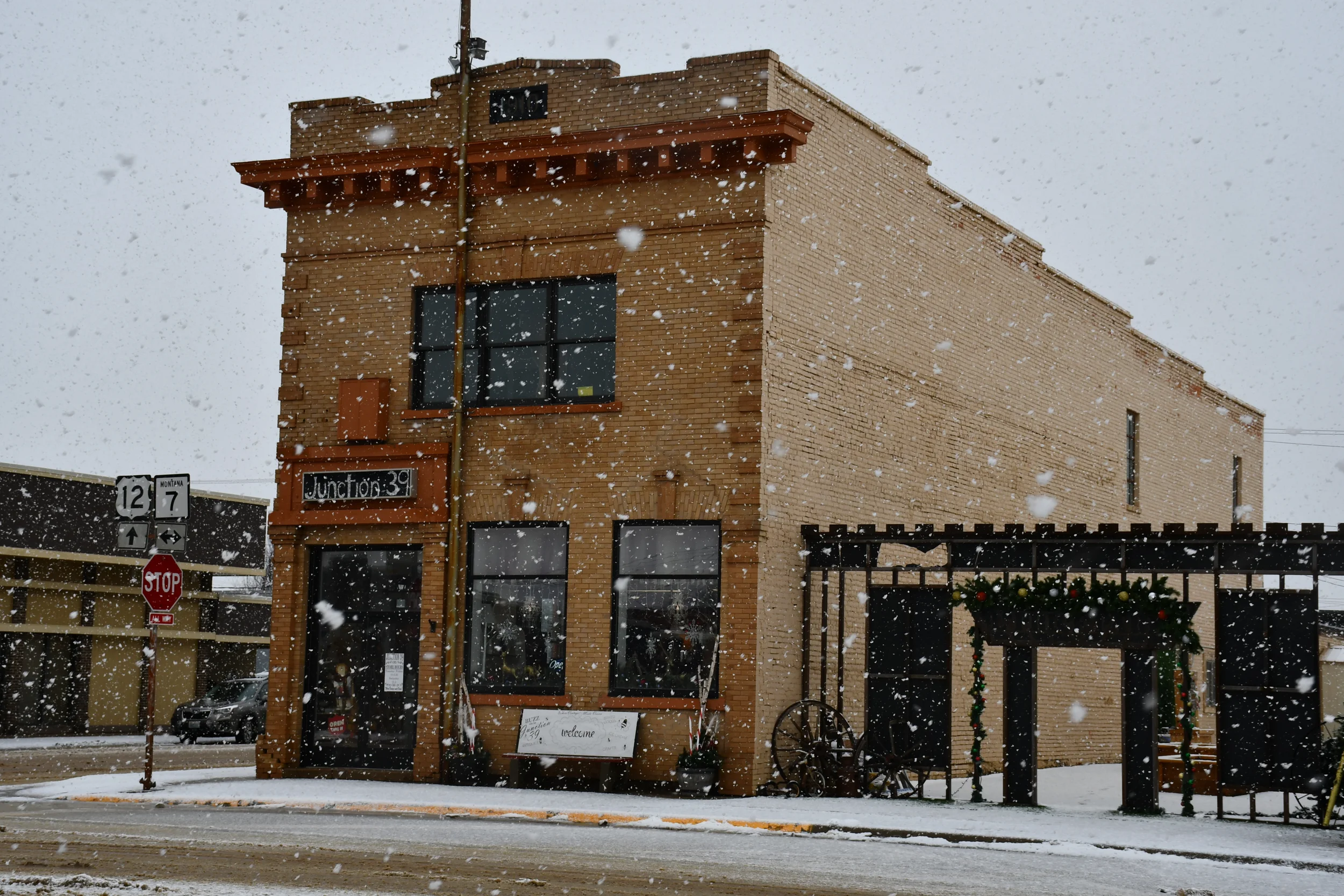 A brick building with a sign that says Junction 39, decorated for the holidays, with snow falling on the street and sidewalk, and a stop sign nearby.