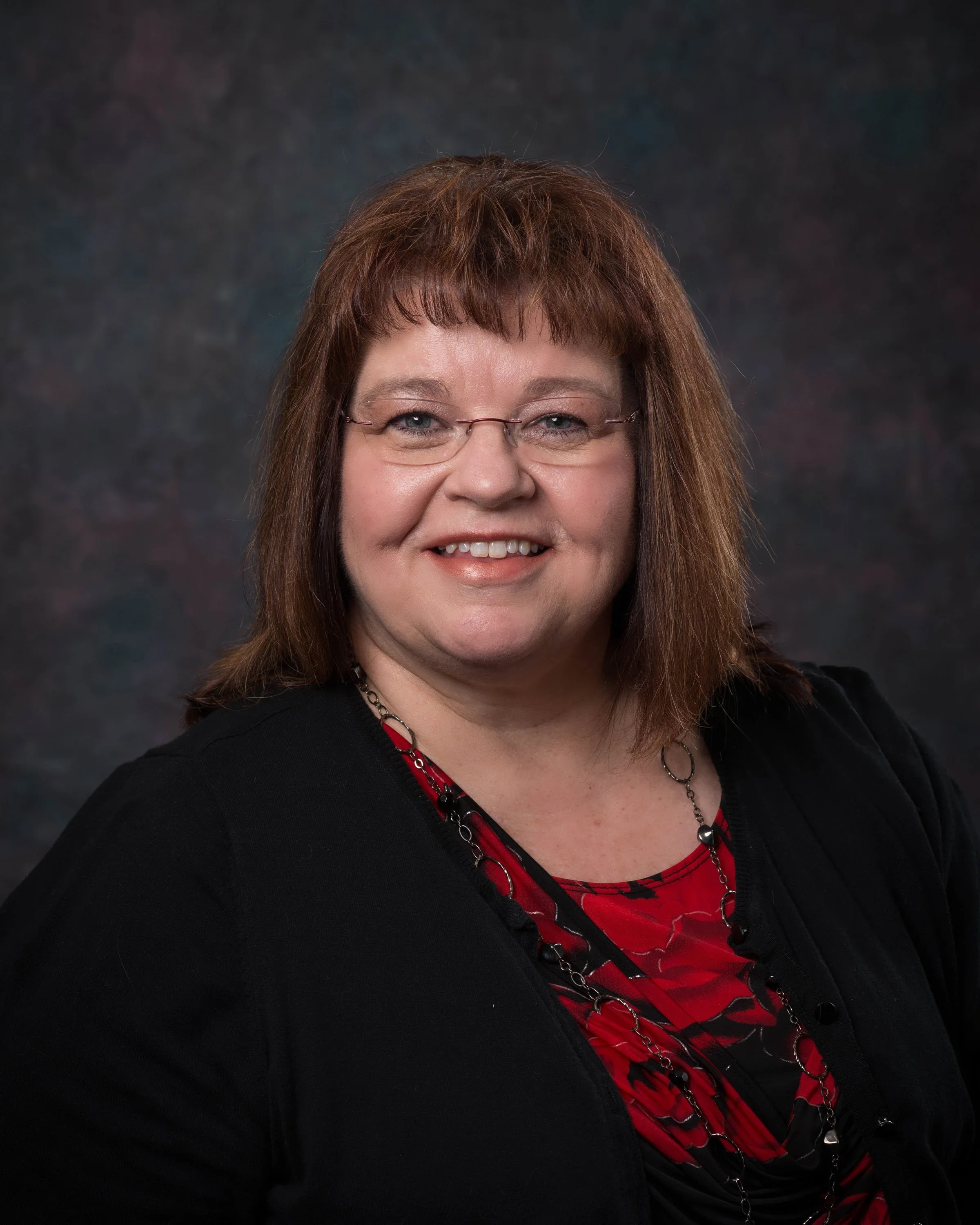 Karol Zachmann with shoulder-length reddish-brown hair, glasses, and a friendly smile, wearing a black jacket and a red patterned blouse, posed against a dark, textured background.
