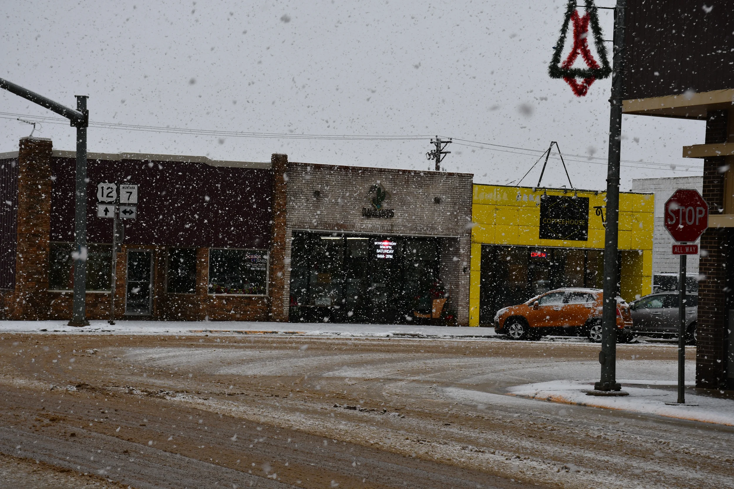 A snowy street scene with a brick building complex featuring a health clinic and a yellow coffeehouse, cars parked on the side, a stop sign, and snow falling heavily.