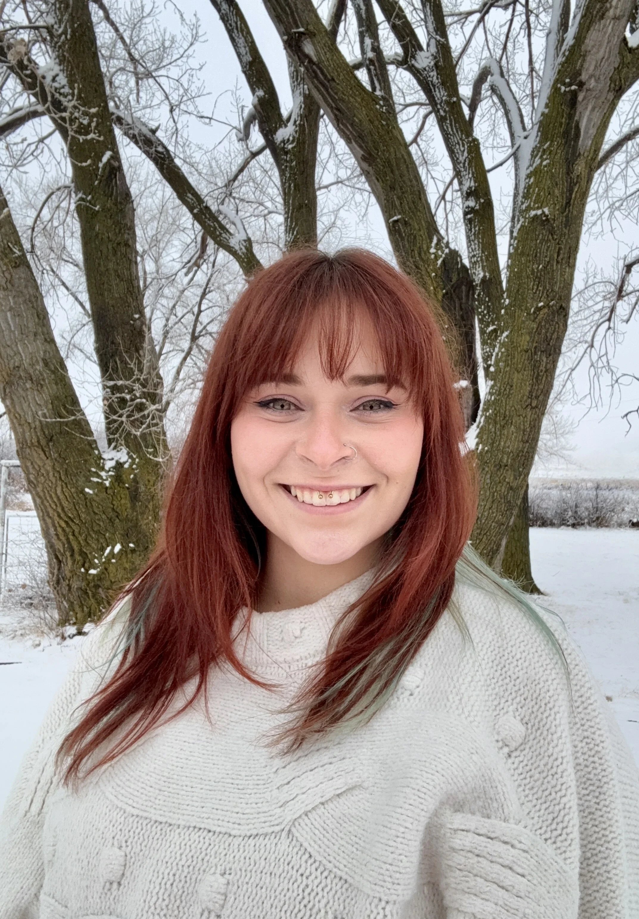 Shelby Klus with red hair and smiling outdoors in front of a snow-covered tree in winter.
