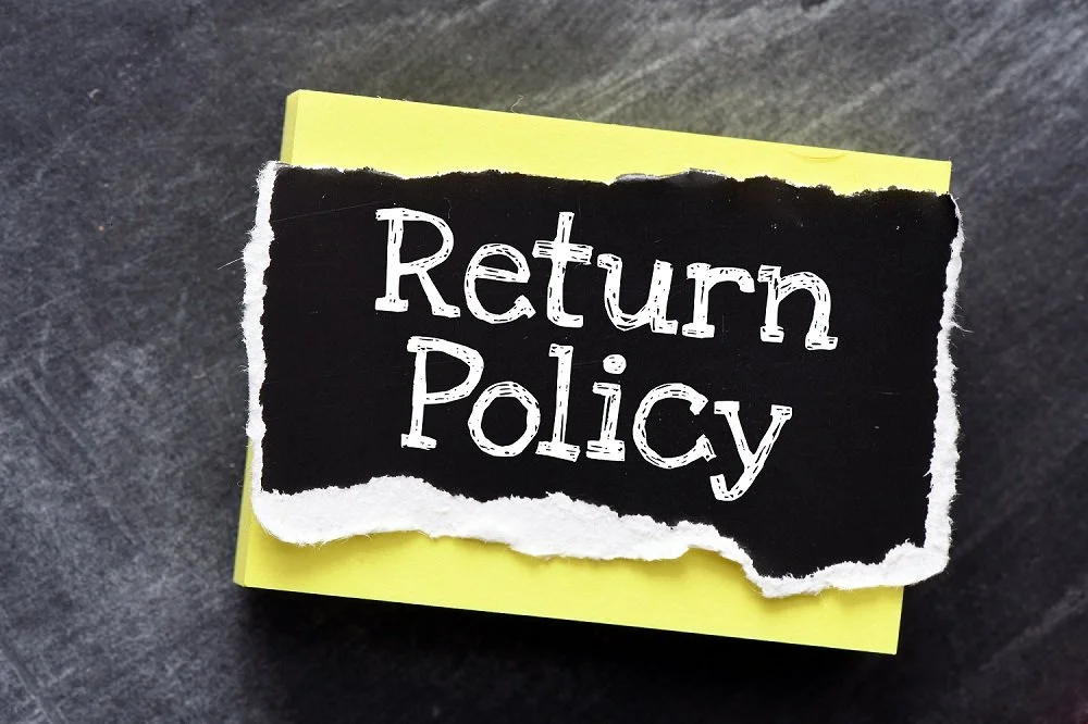 A torn black paper with white text reading 'Return Policy' on a yellow sticky note, placed on a dark textured surface.
