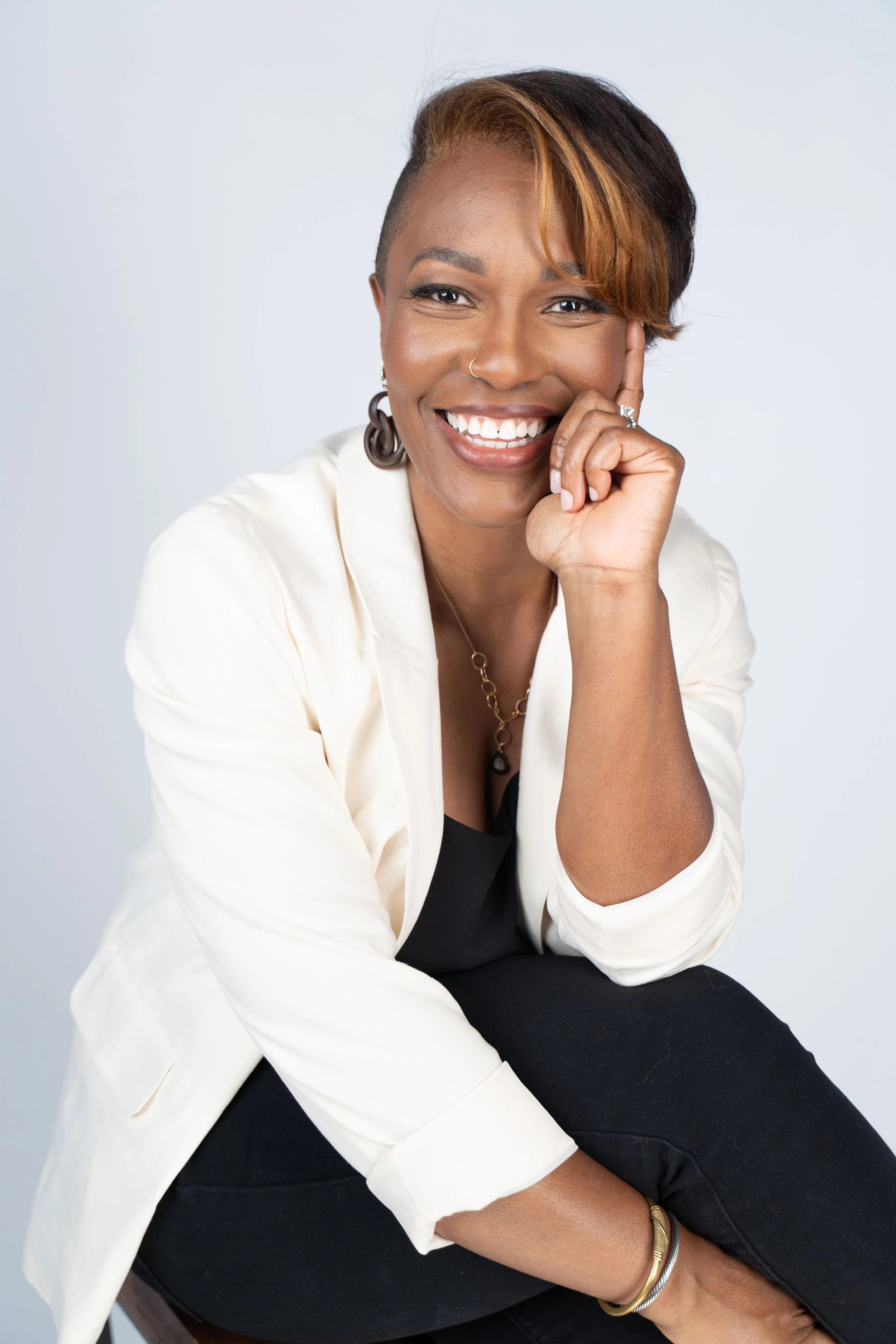 A smiling woman with short, styled hair, wearing a white blazer over a black top, gold jewelry, and sitting in a relaxed pose against a plain background.