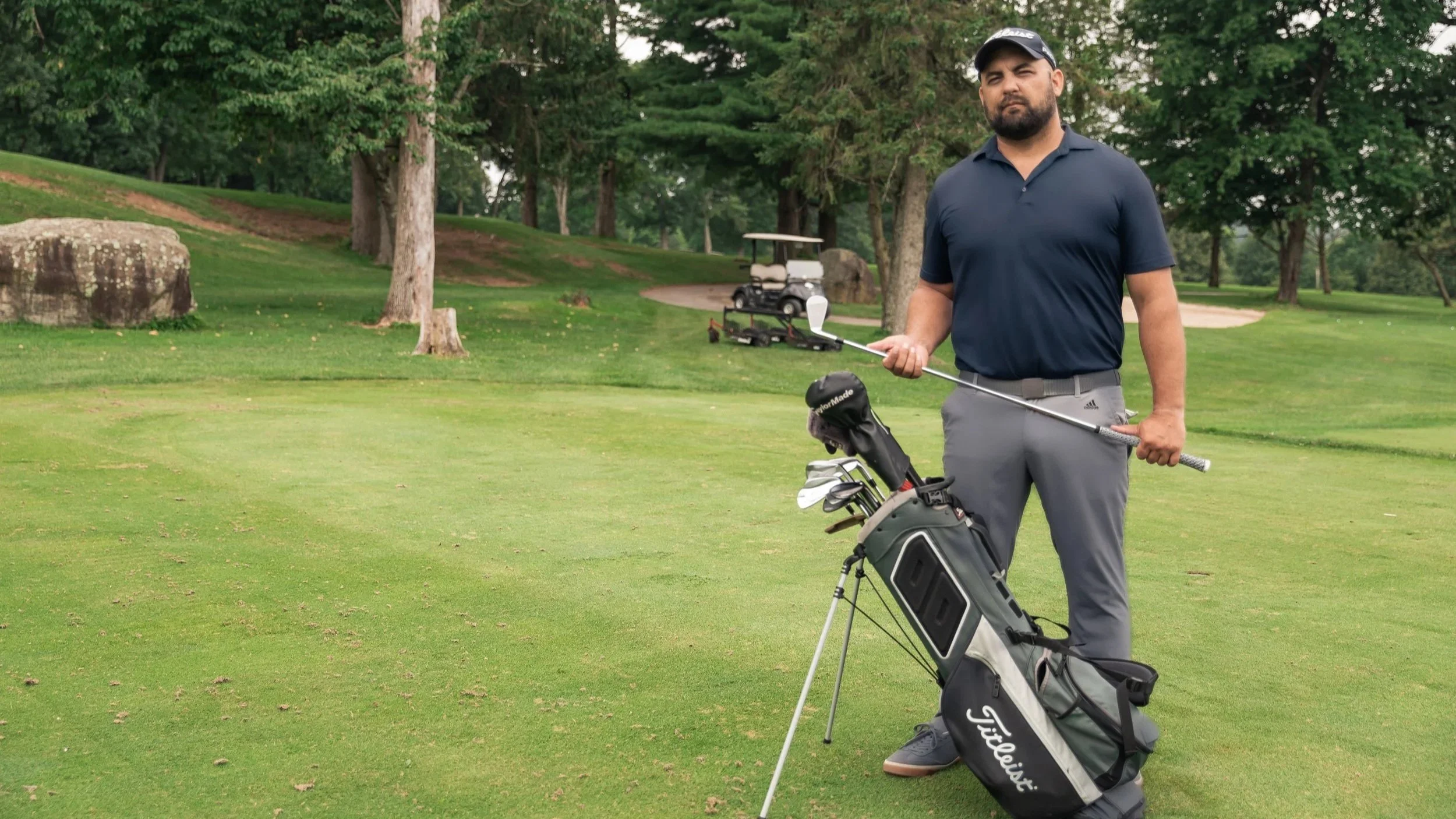 A man standing on a golf course with a golf club and a golf bag, surrounded by trees and grass, with golf carts in the background.