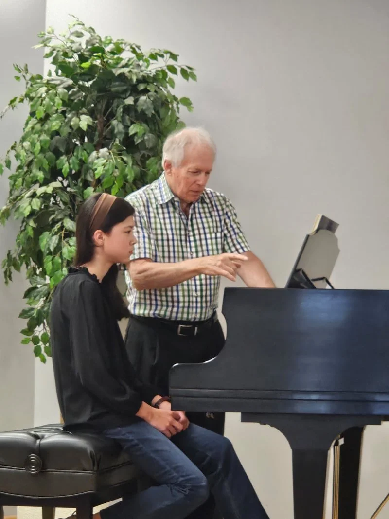 An elderly man teaching a young girl how to play the piano in a music room with a large green plant in the background.