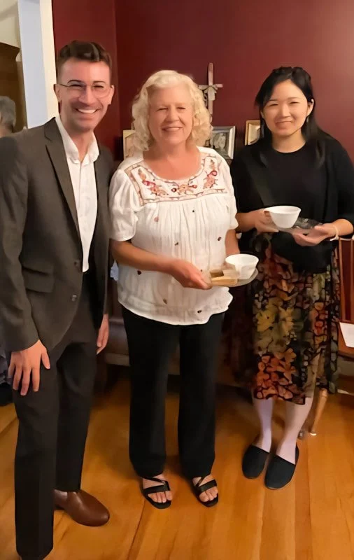 Three people standing indoors; a man in a suit, a woman in a white blouse, and a woman in a black top and floral skirt, all smiling and holding bowls.