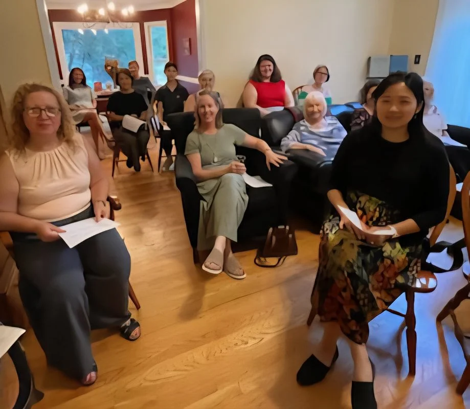 Group of women and men seated in a living room, some holding papers, attending a meeting or gathering.