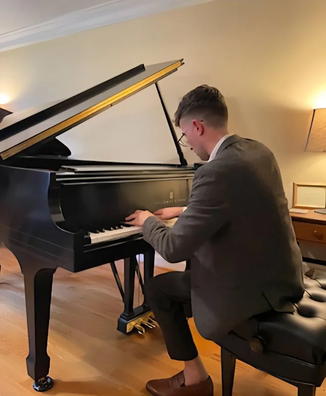 Young man in grey blazer and glasses playing grand piano in a cozy, warmly-lit room.