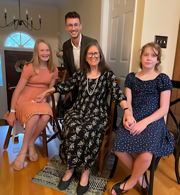 A family of four, including two women and two young girls, smiling and posing together in a home dining area.