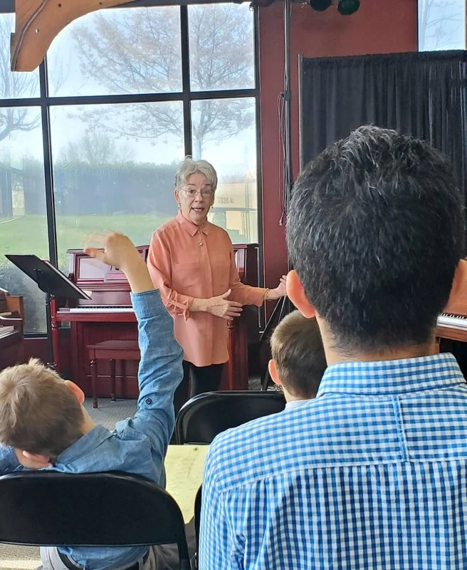 An older woman with short gray hair and glasses speaking in front of a group of children and adults in a room with large windows, a piano, and a black curtain.
