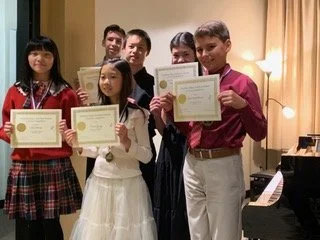 Five young students holding certificates, standing together in a room with yellow curtains and a piano in the background, celebrating their achievement.