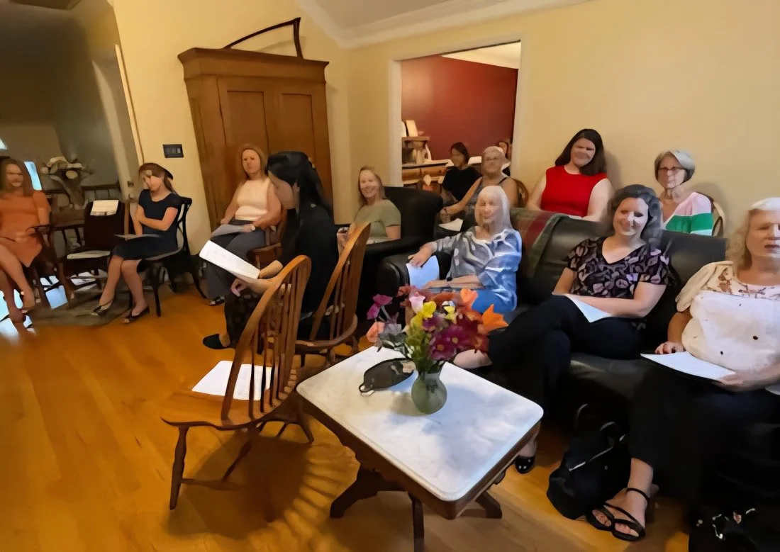 A group of women sitting in a living room, some holding papers, with a table with flowers in front of them.