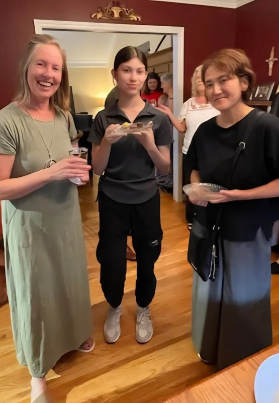 Three women standing indoors, two holding small plates of food, smiling at the camera. The woman on the left wears a light green dress, the woman in the middle wears a black uniform, and the woman on the right wears a black top and gray skirt. There 