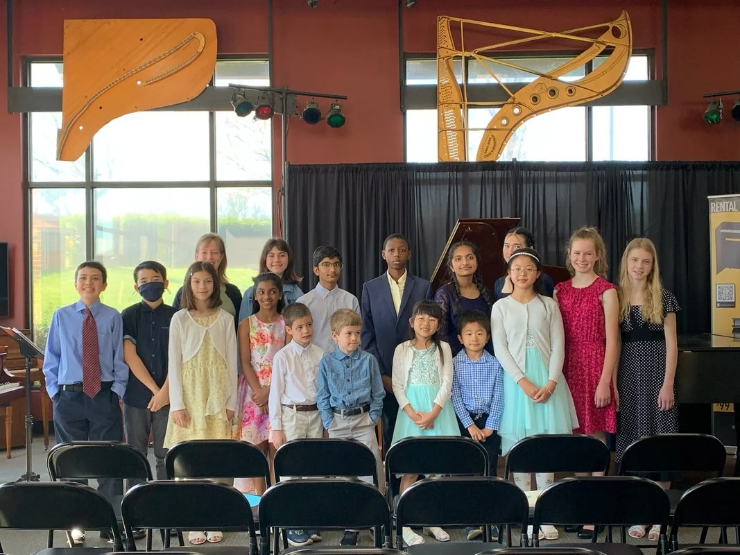 Group of children dressed in formal and semi-formal attire standing on a stage with a black curtain, piano, and large wooden musical note sculptures in the background, likely after a musical performance or recital.