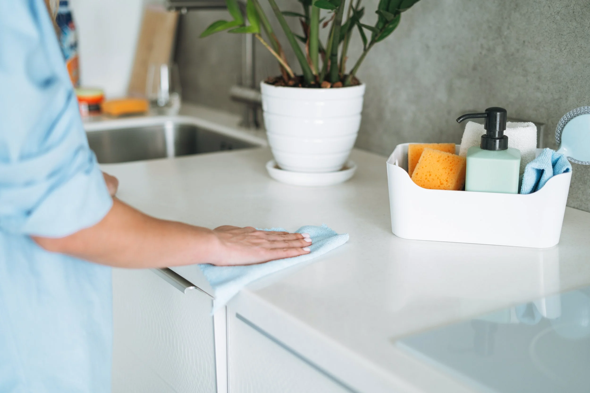 Person wiping a white kitchen countertop with a blue cloth. There is a white container holding cleaning supplies, sponges, a pump bottle, and cloths. A potted plant is in the background beside a sink.
