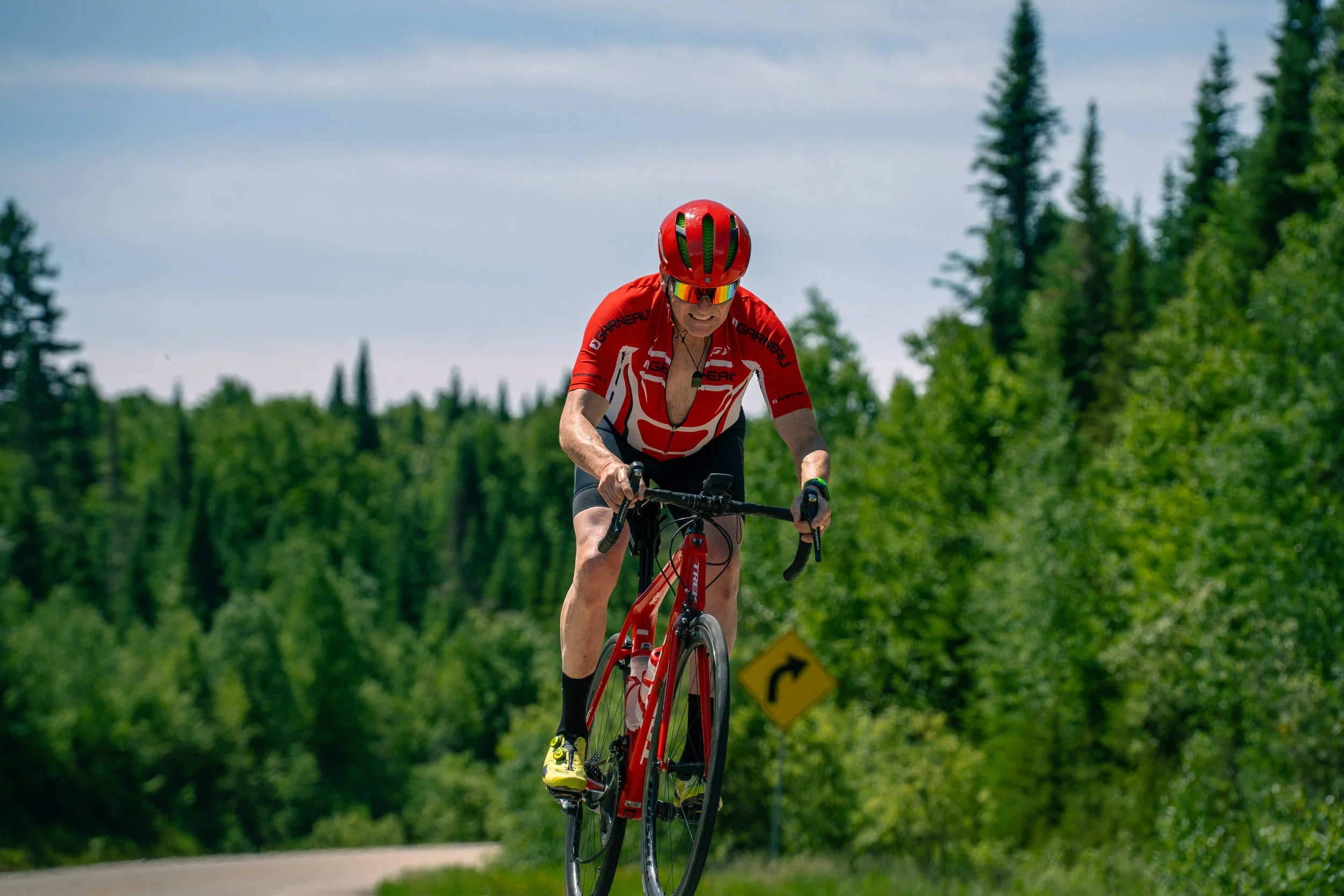 Un homme âgé faisant du vélo sur une route forestière, portant un casque rouge, un maillot rouge et noir, des lunettes de soleil et des chaussures jaunes. La route est bordée d'arbres verts et un panneau de signalisation indique un virage à droite.