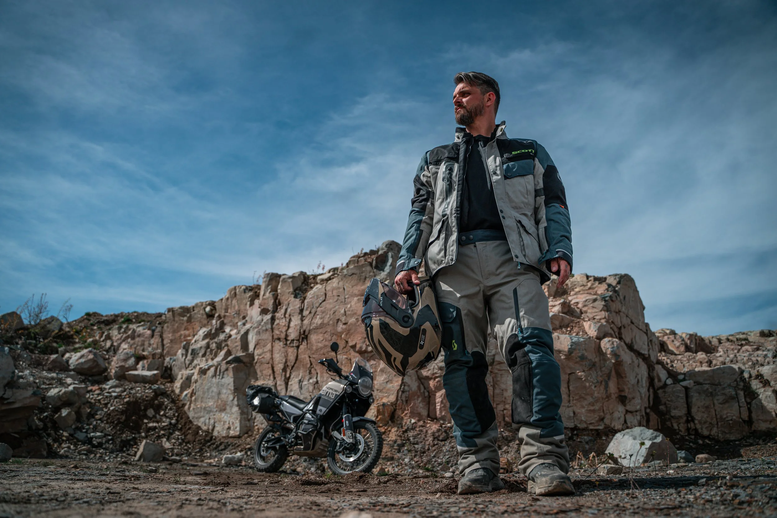 Un homme en équipement de moto tout-terrain avec un casque à la main, debout dans un paysage rocheux avec une moto à l'arrière.