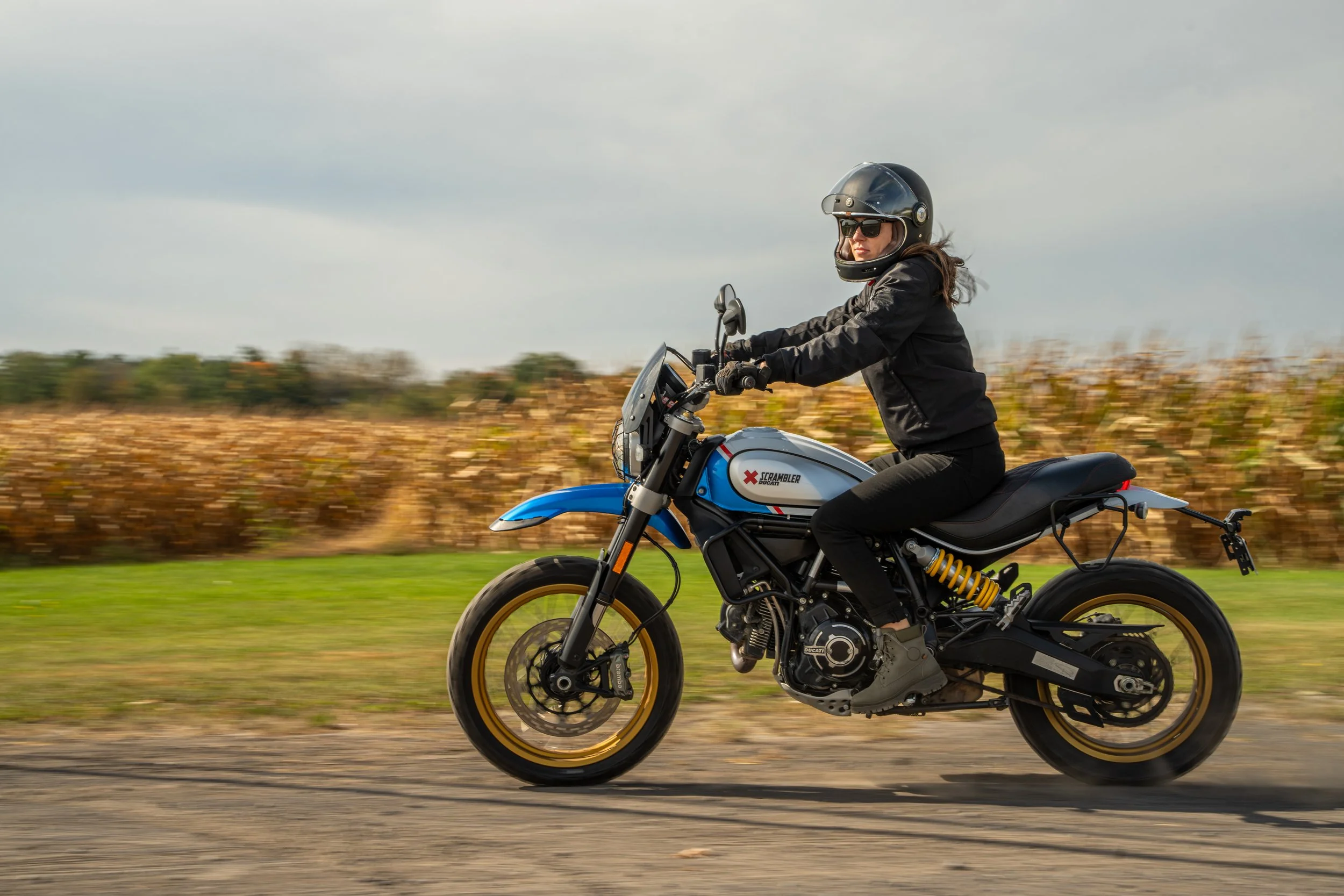 Une femme portant un casque et un vêtement noir monte une moto de couleur blanche et bleue, roulant sur une route avec un champ en arrière-plan.