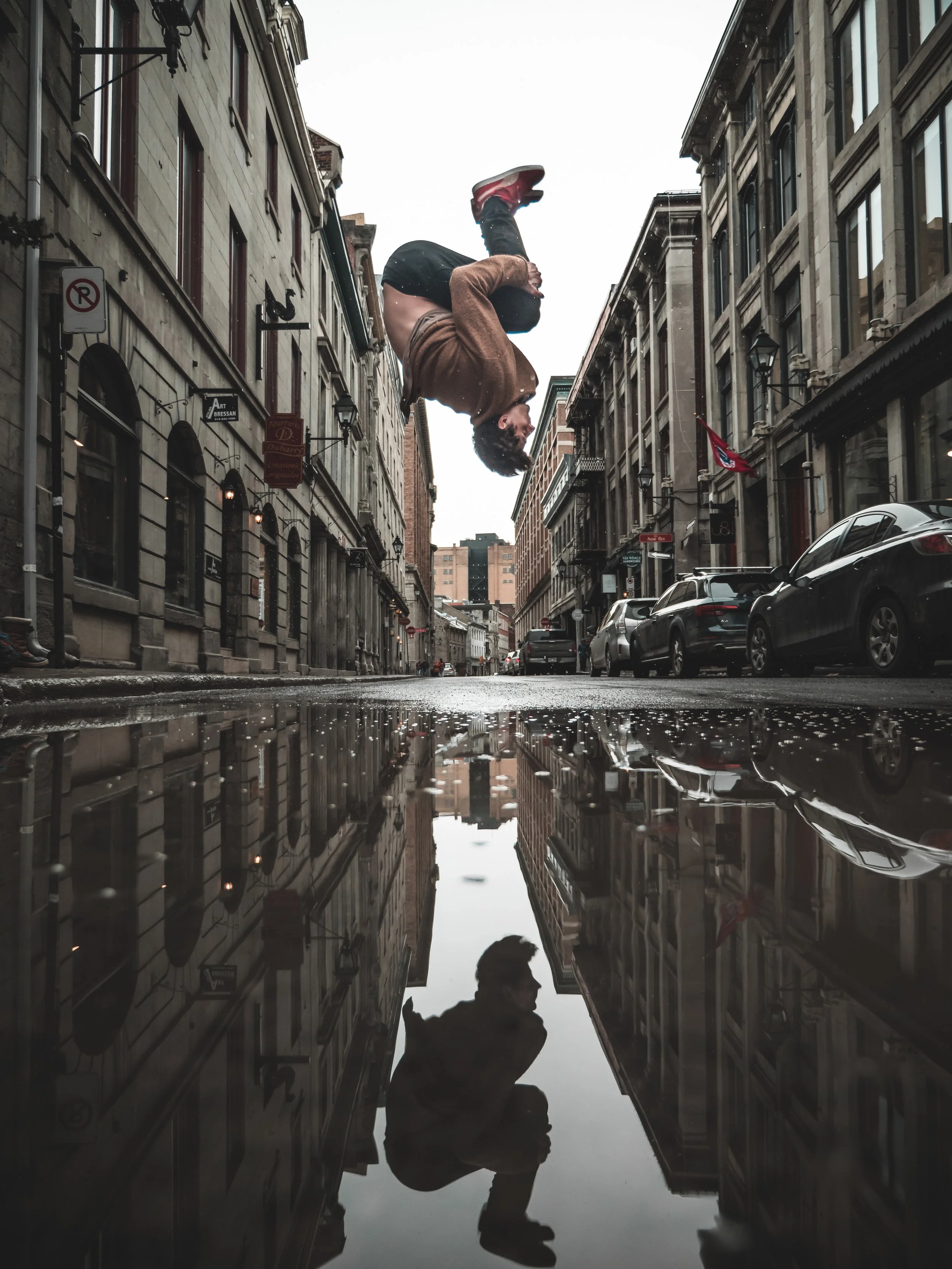 Une rue urbaine avec des bâtiments anciens, des voitures garées et un homme qui saute ou tombe, son reflet visible dans une flaque d'eau au sol.