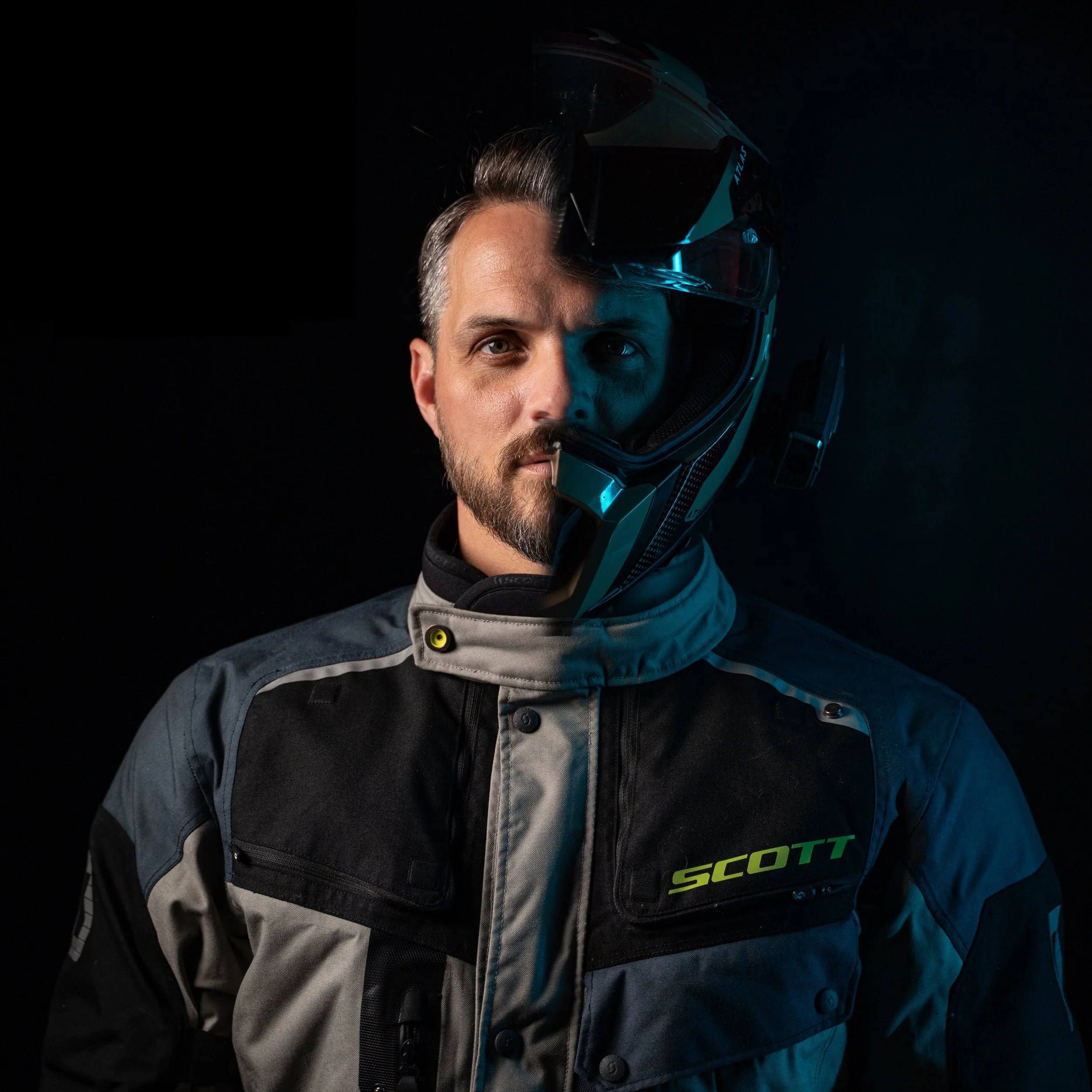 Homme portant une combinaison de moto et un casque partiellement levé, face à un fond noir.