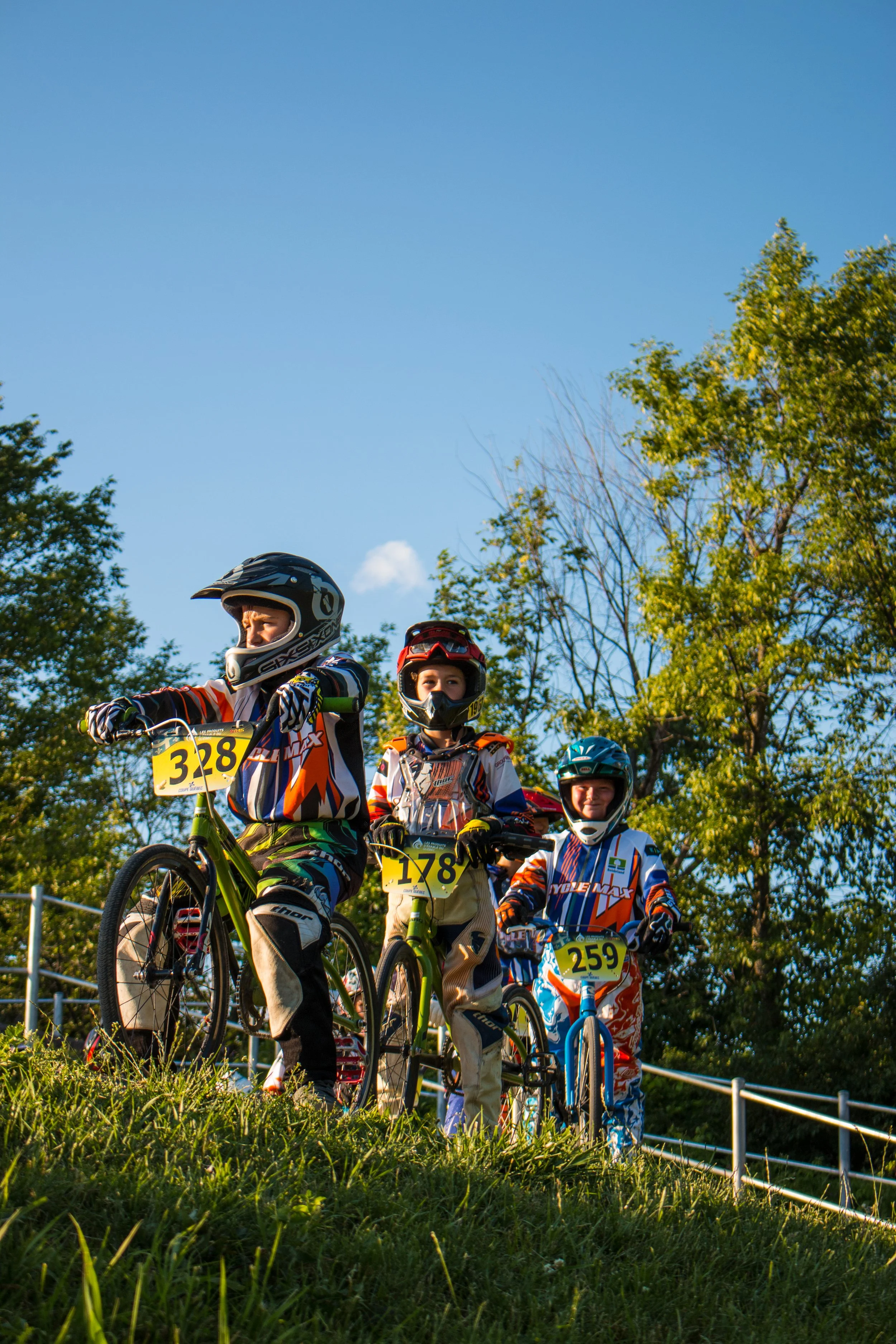 Trois jeunes cyclistes en vêtements de motocross, portant casques, positionnés sur une pente herbeuse, avec un ciel clair et des arbres en arrière-plan.