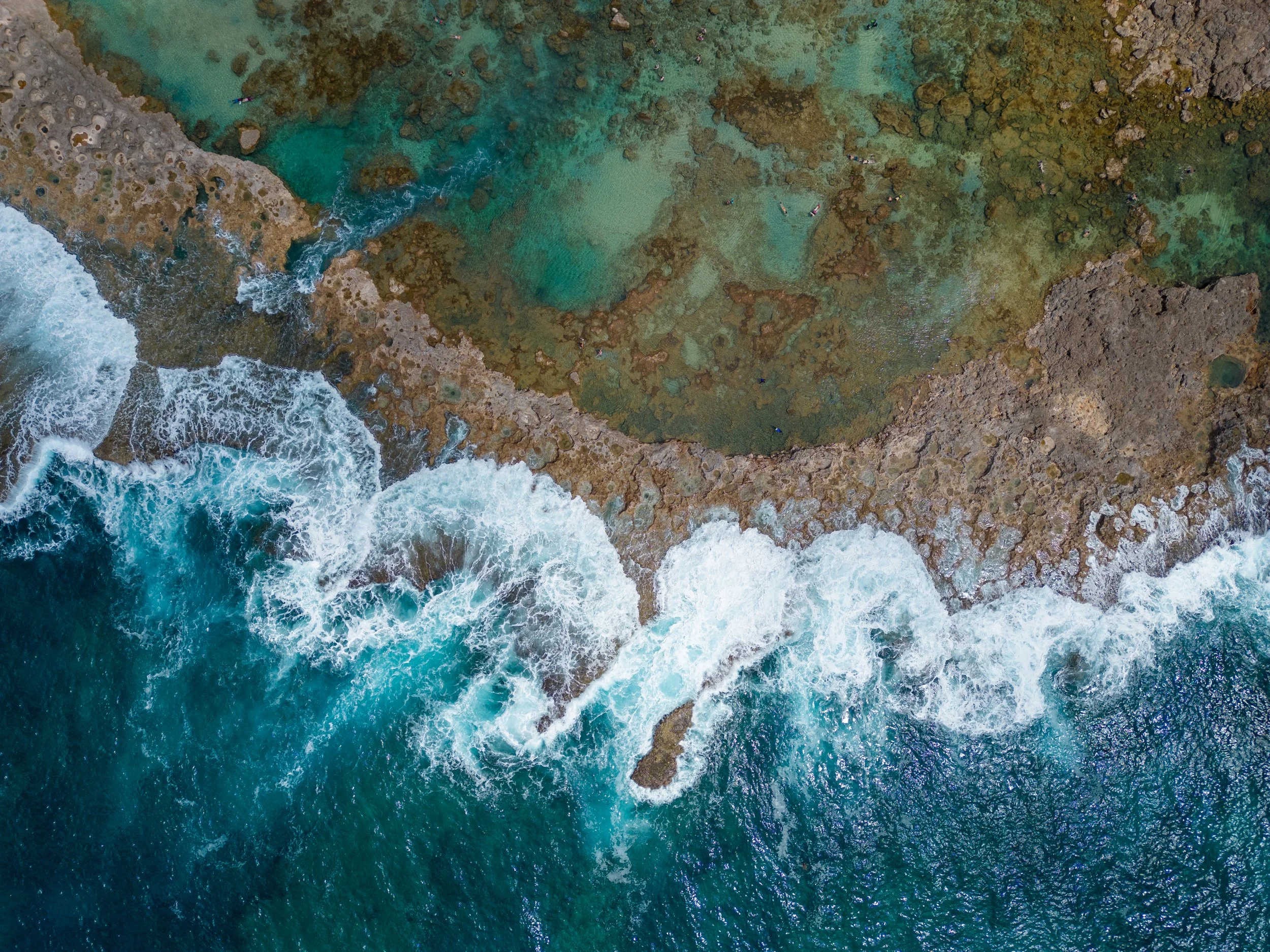 Vue aérienne d'une côte rocheuse avec des vagues s'écrasant contre la roche et une zone d'eau peu profonde avec des teintes vertes et turquoises.
