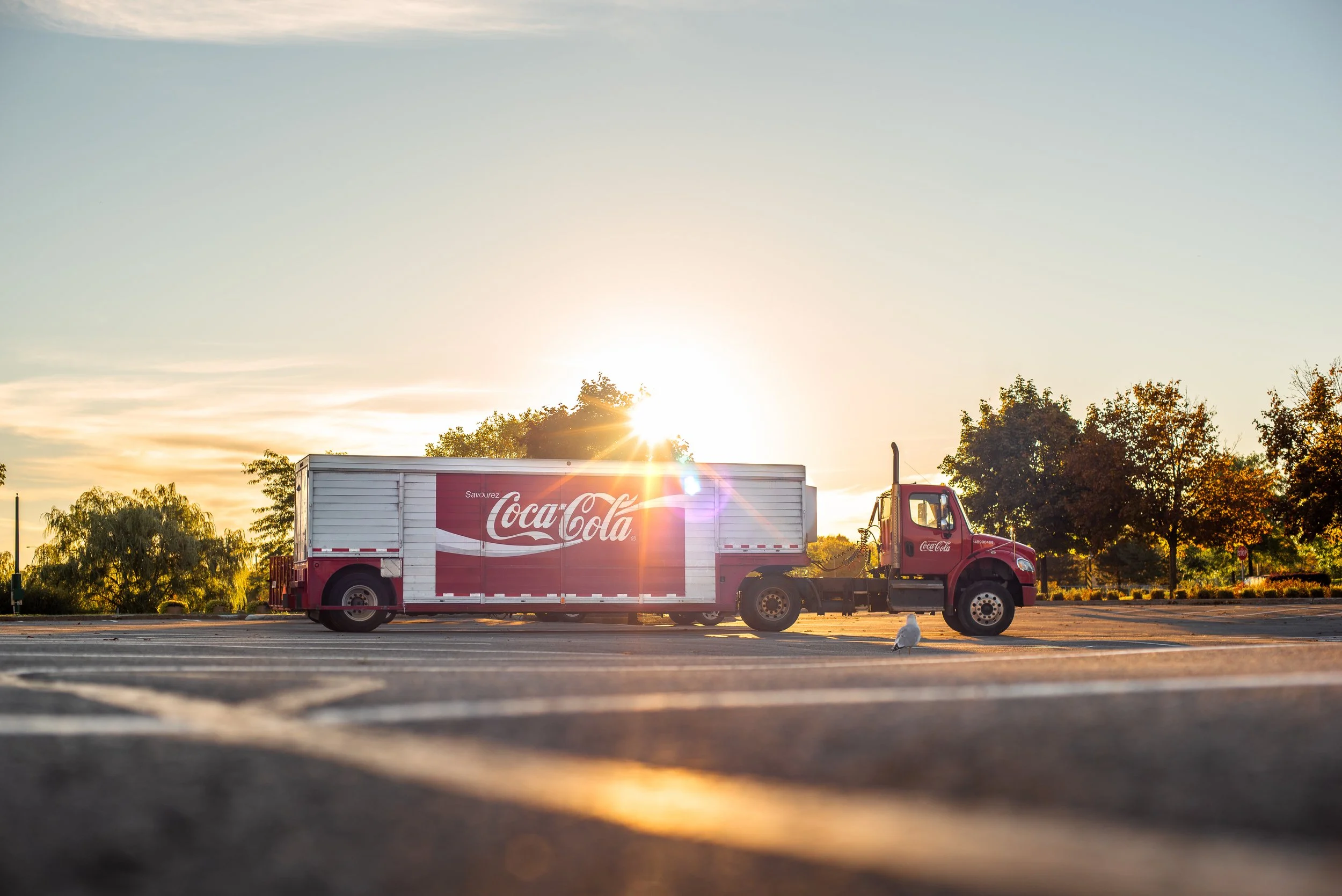 Camion de livraison de Coca-Cola stationné dans un parking avec le soleil couchant en arrière-plan, entouré d'arbres.
