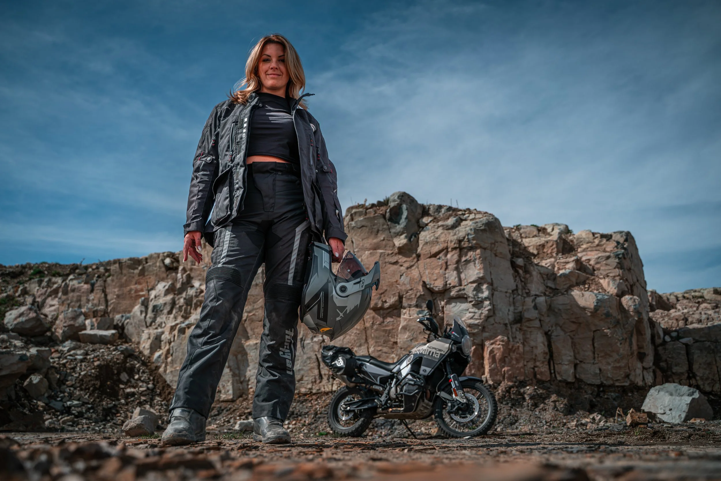 Femme motarde en équipement de moto tout-terrain, tenant un casque, devant une moto dans un paysage rocheux avec un ciel dégagé.