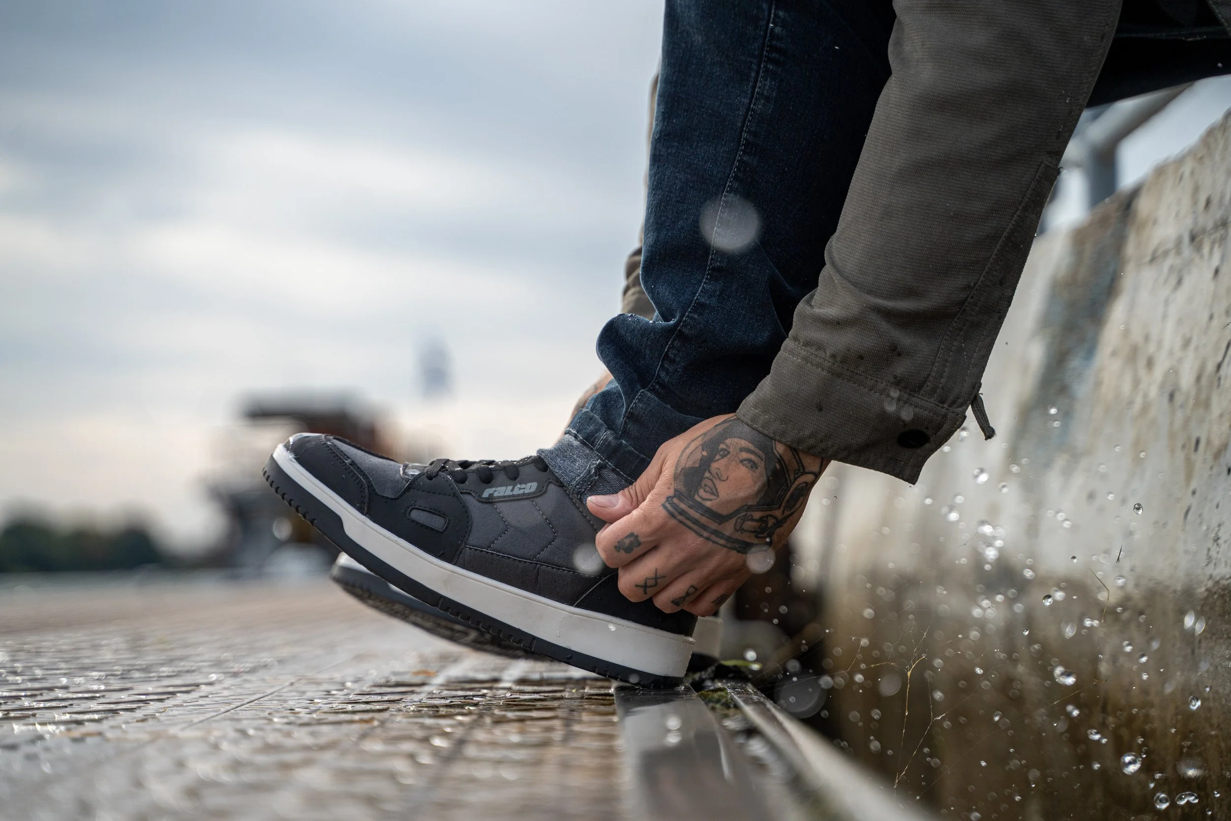 Une personne portant des chaussures de sport noires et des vêtements décontractés, assise sur un quai en bois, ajustant sa chaussure, avec des gouttes d'eau sur le sol et la pluie tombant dans un ciel nuageux.