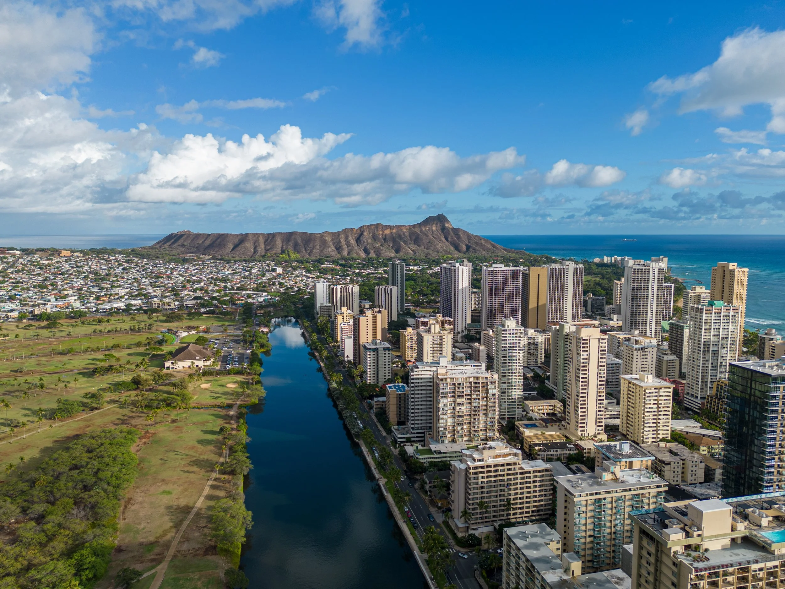 Vue aérienne d'une ville côtière avec gratte-ciel, un fleuve, des montagnes et l'océan en arrière-plan, sous un ciel partiellement nuageux.