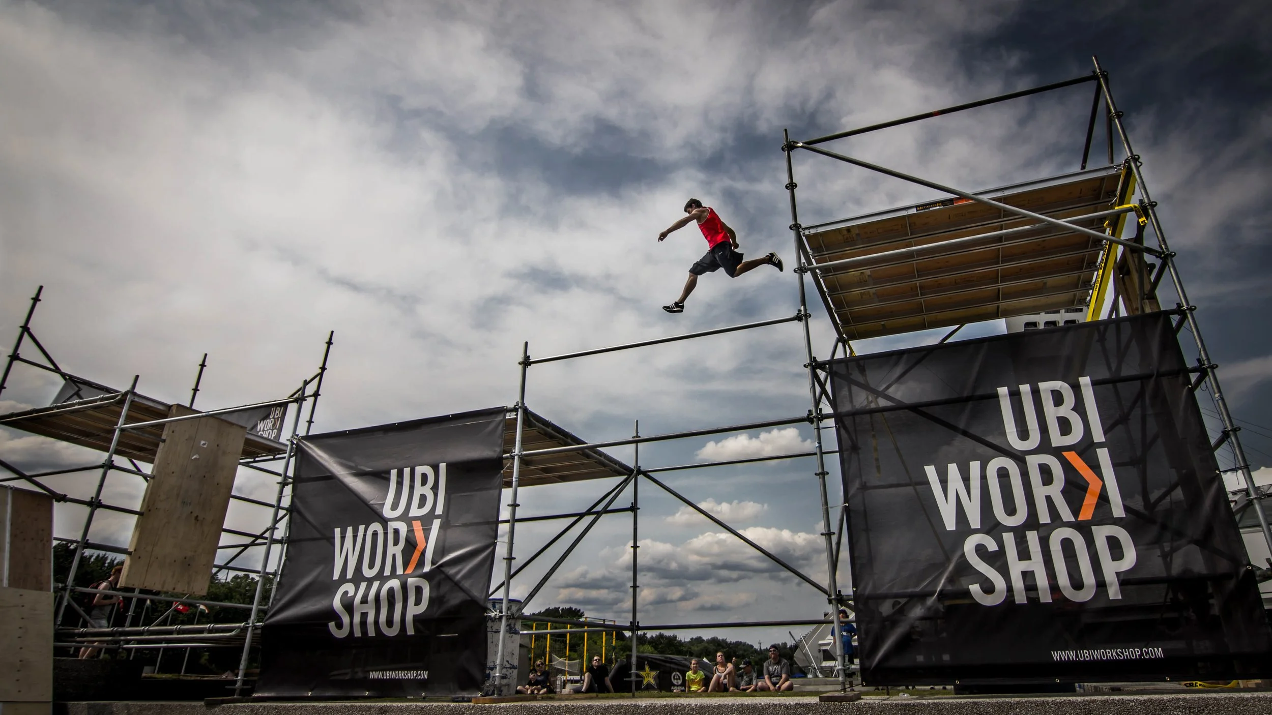 Un homme en salopette rouge sautant d'une plateforme d'escalade en scaffolding lors d'un événement UBI WORKSHOP, avec un groupe de spectateurs en dessous et un ciel nuageux en arrière-plan.